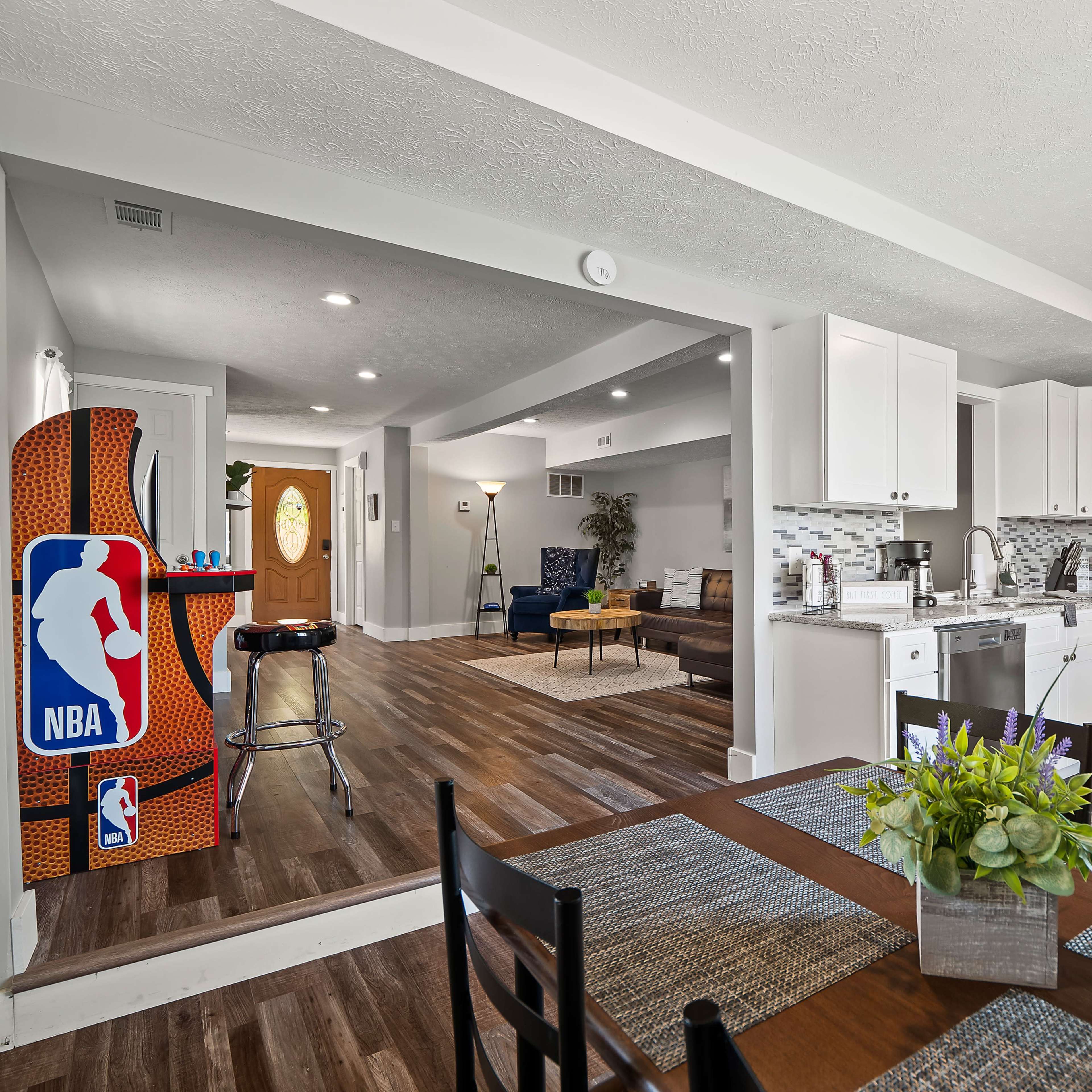 A modern kitchen and dining area with white cabinetry, stainless steel appliances, and a view into a living room featuring an NBA-themed arcade game.