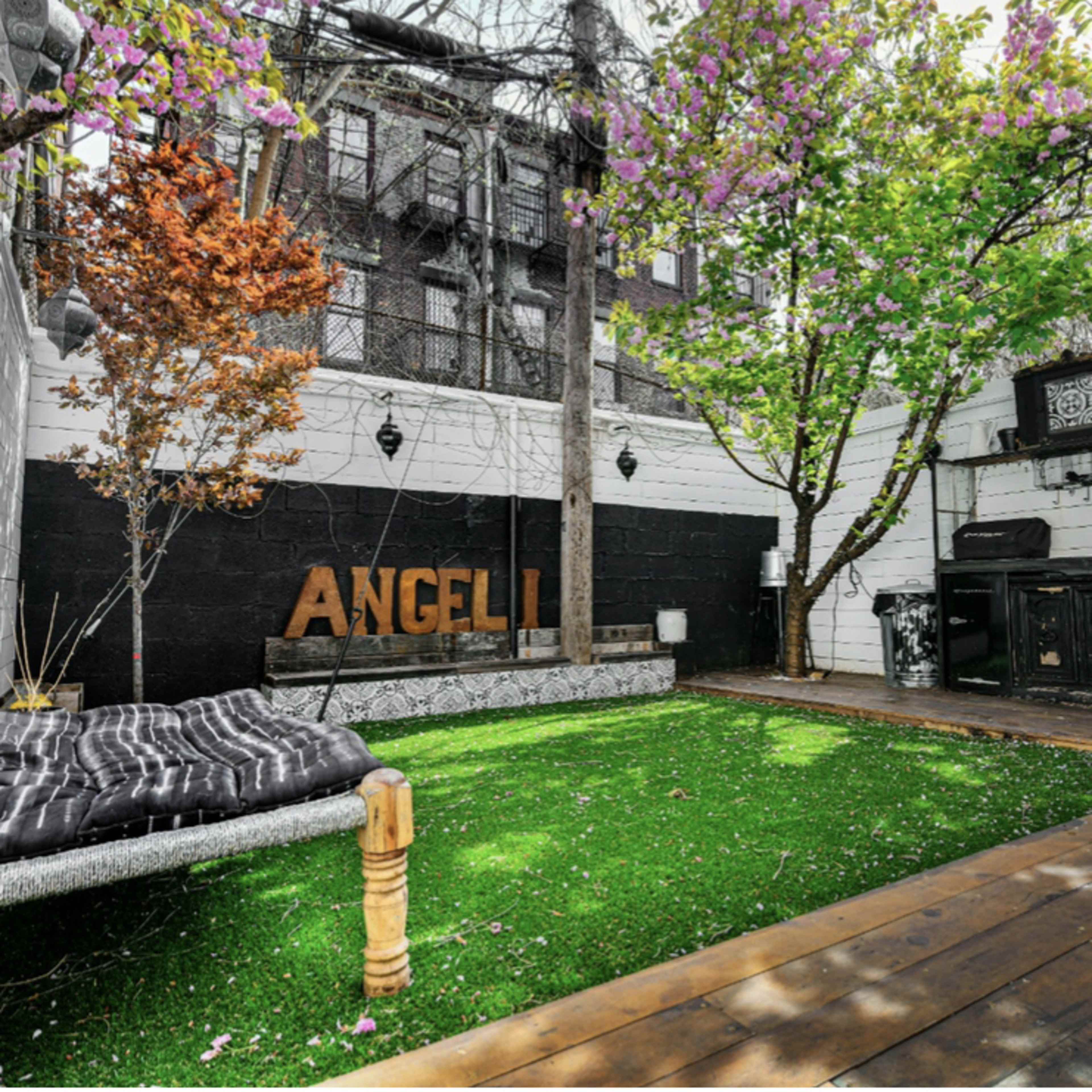 A landscaped outdoor space featuring a grassy area, a wooden deck, a bench, and a wall with the word "ANGELI" displayed.