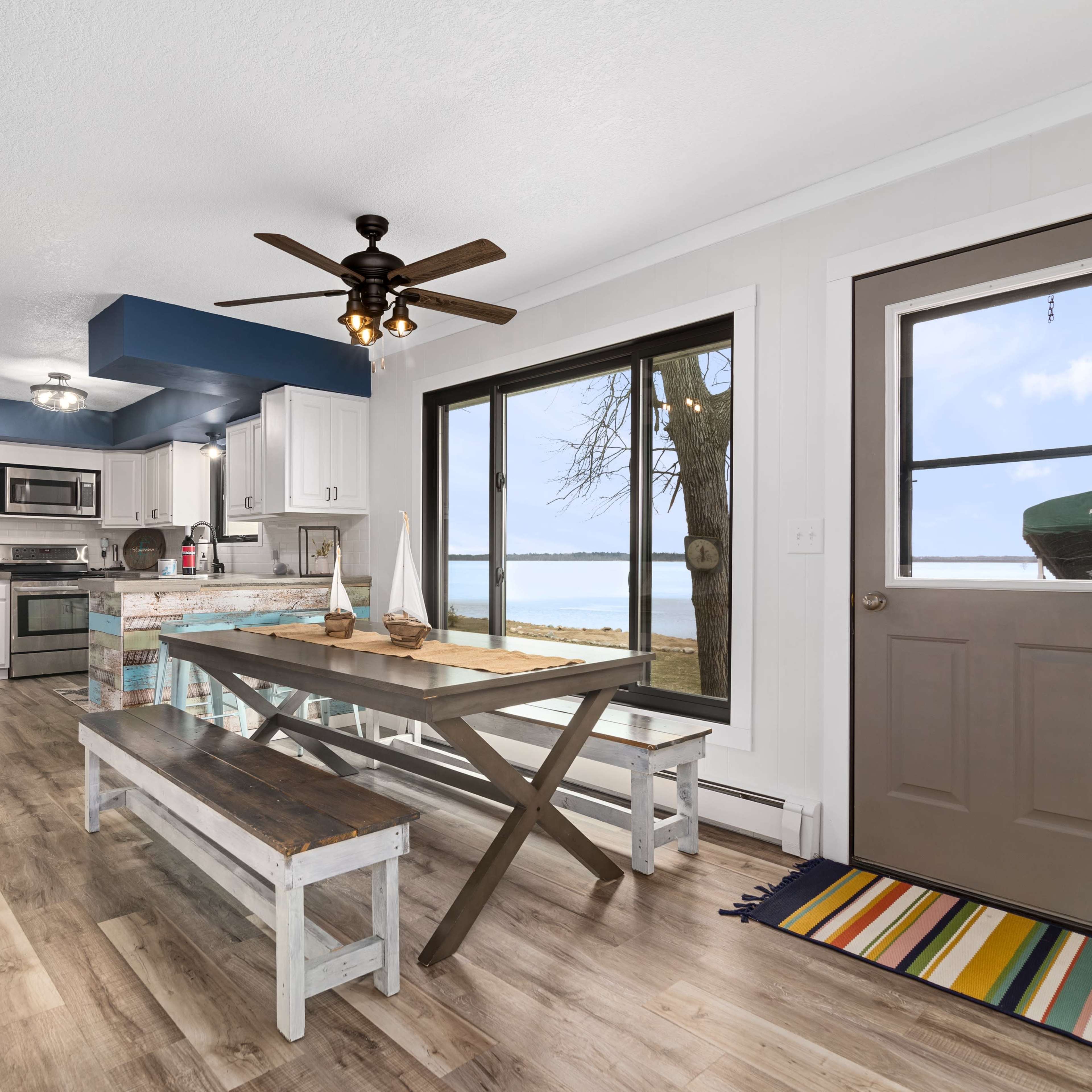 A light-filled kitchen and dining area with a large window overlooking a body of water, featuring a table with benches and a brown front door.