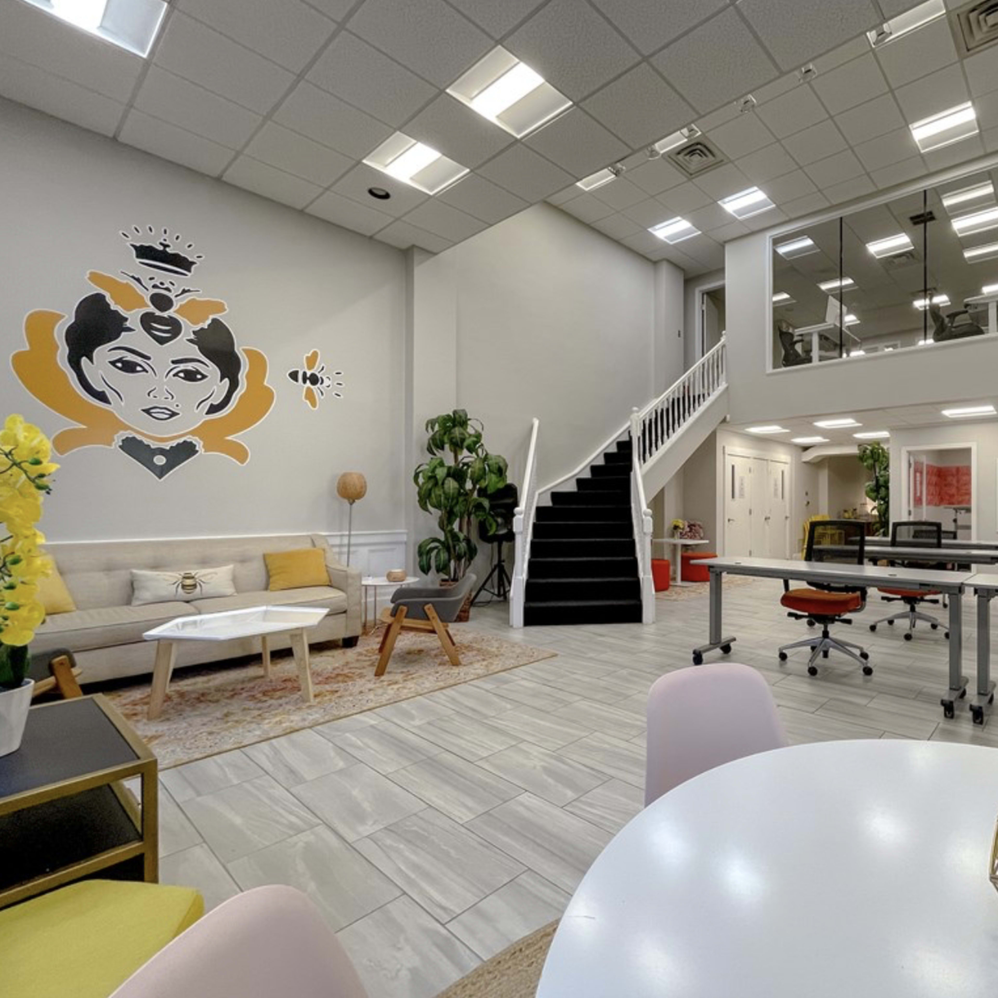 A modern office space features a seating area with a decorative mural, large windows, and an upper-level office with a black staircase.