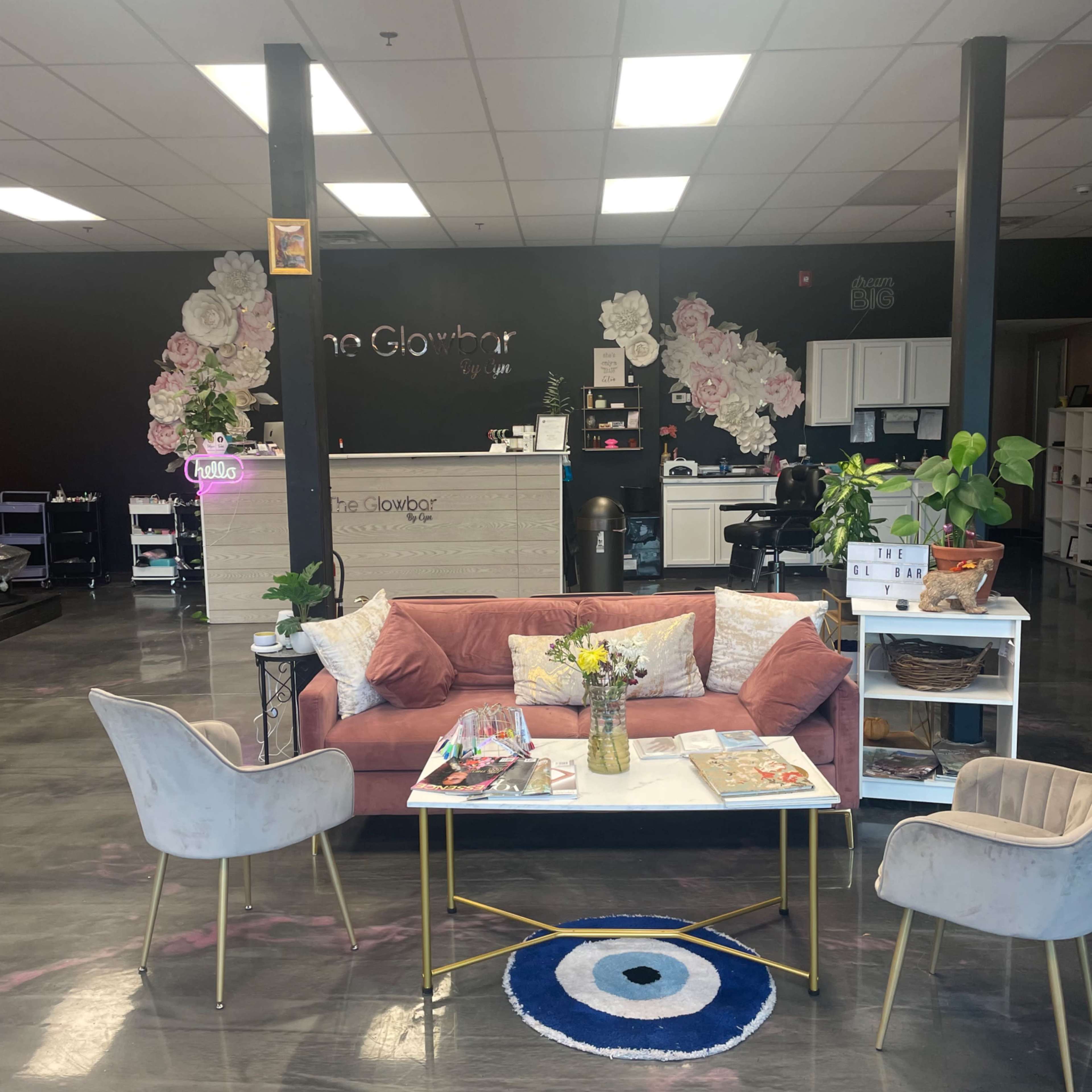 A modern beauty salon reception area with a pink couch, a coffee table, decorative flowers on the wall, and stylish chairs.