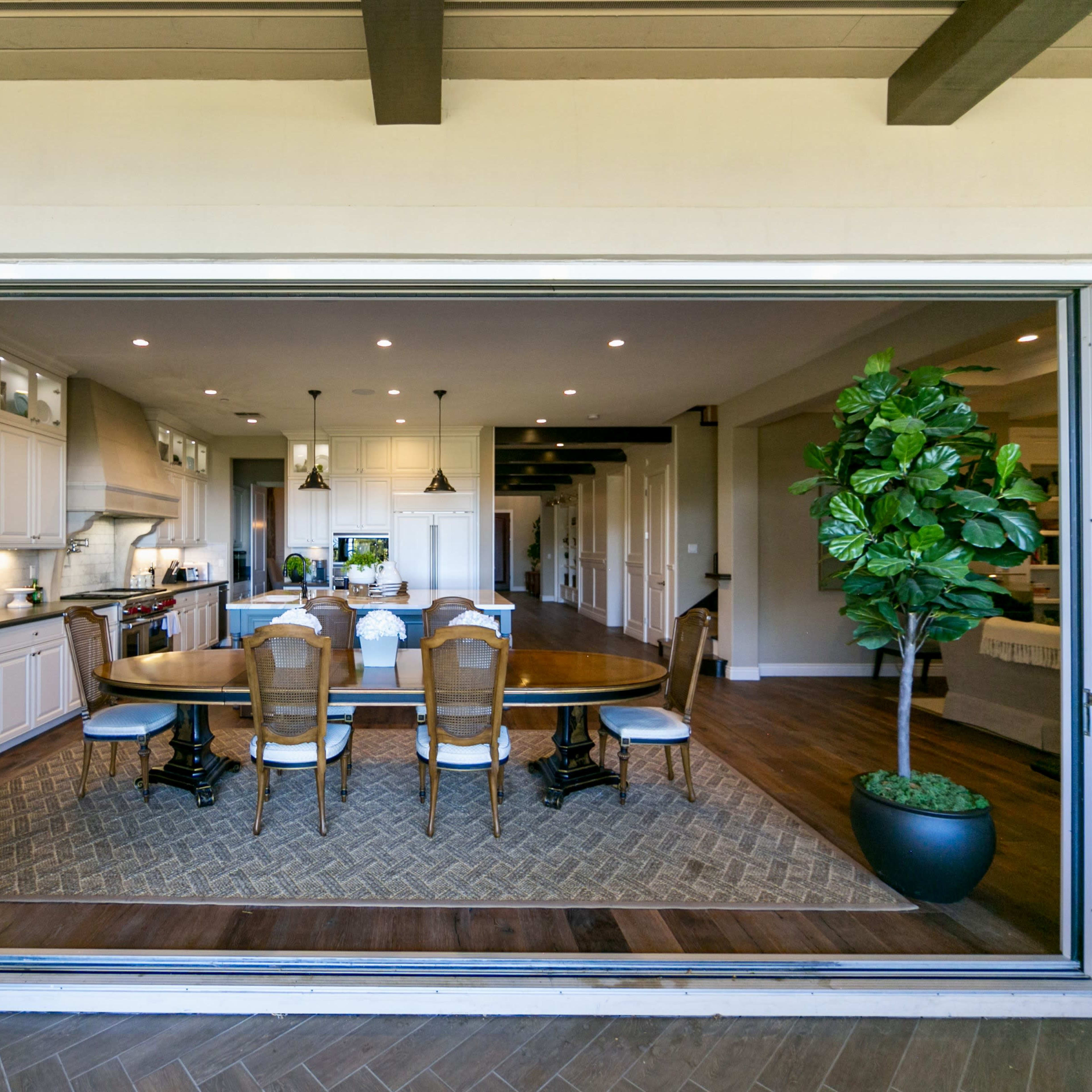 A spacious kitchen and dining area with a large wooden table, stylish chairs, and a potted plant, visible through large sliding glass doors.