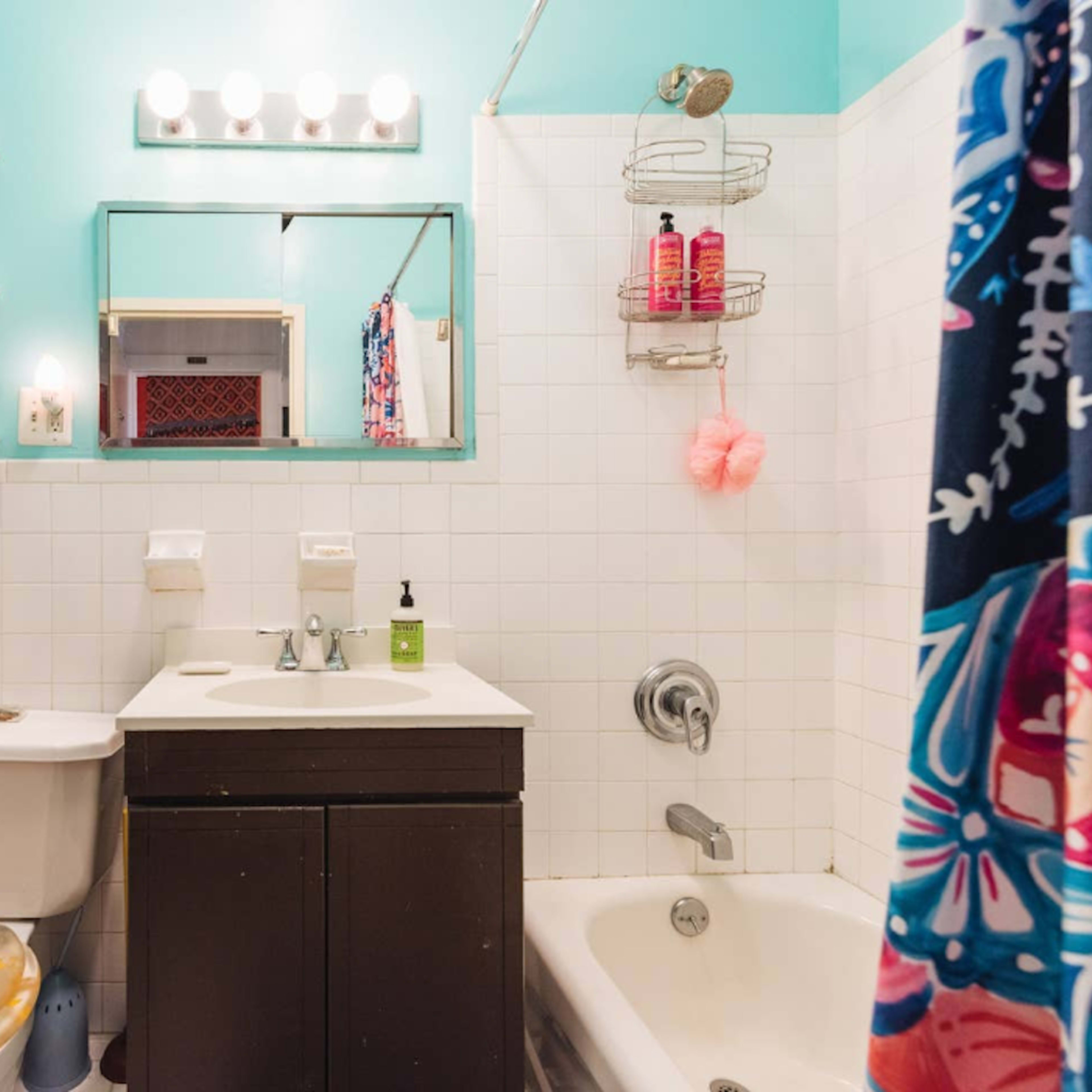 A small bathroom with a turquoise wall, a white tiled bathtub, a dark vanity with a sink, and a shower curtain featuring a colorful pattern.