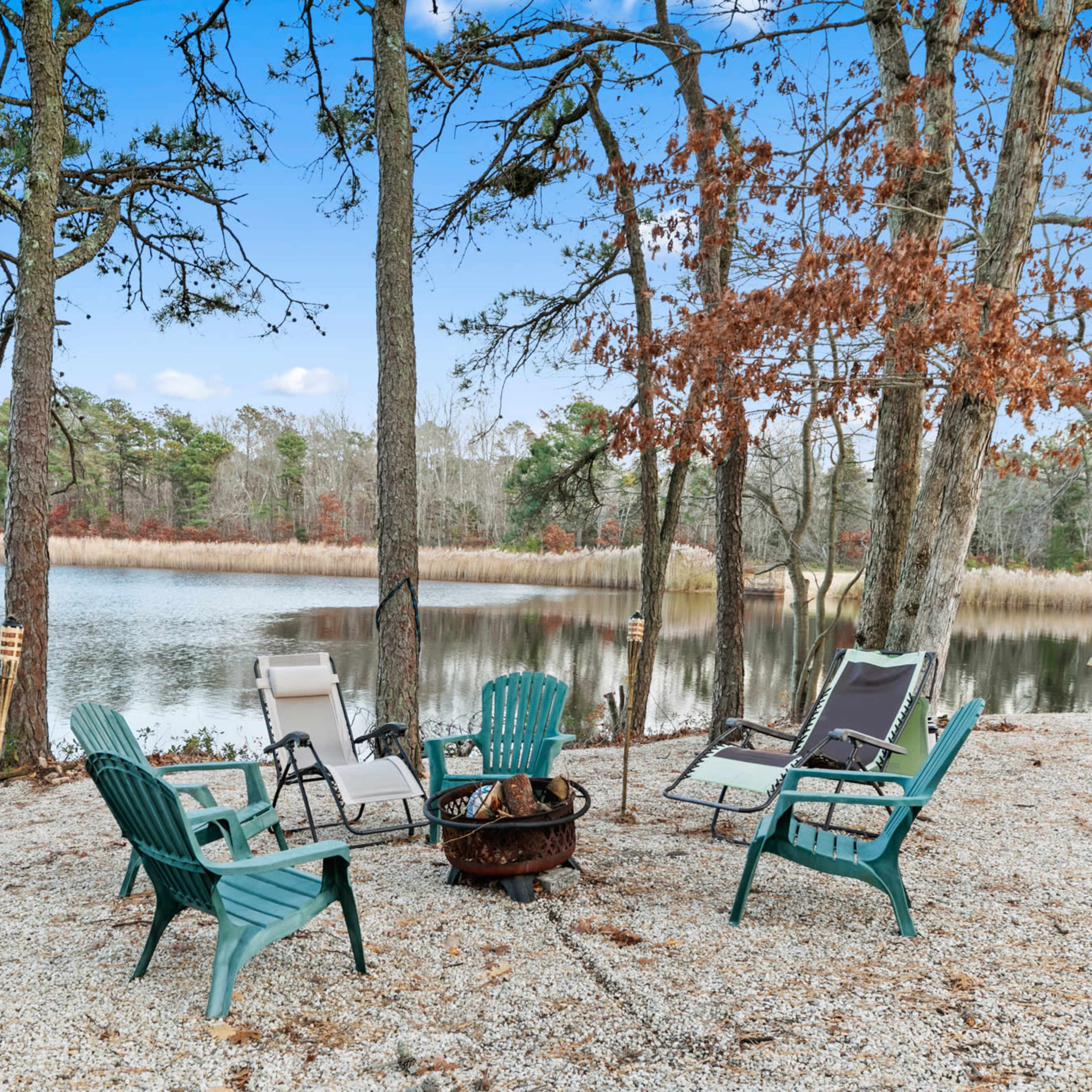 A fire pit surrounded by several chairs near a calm lake, framed by trees.
