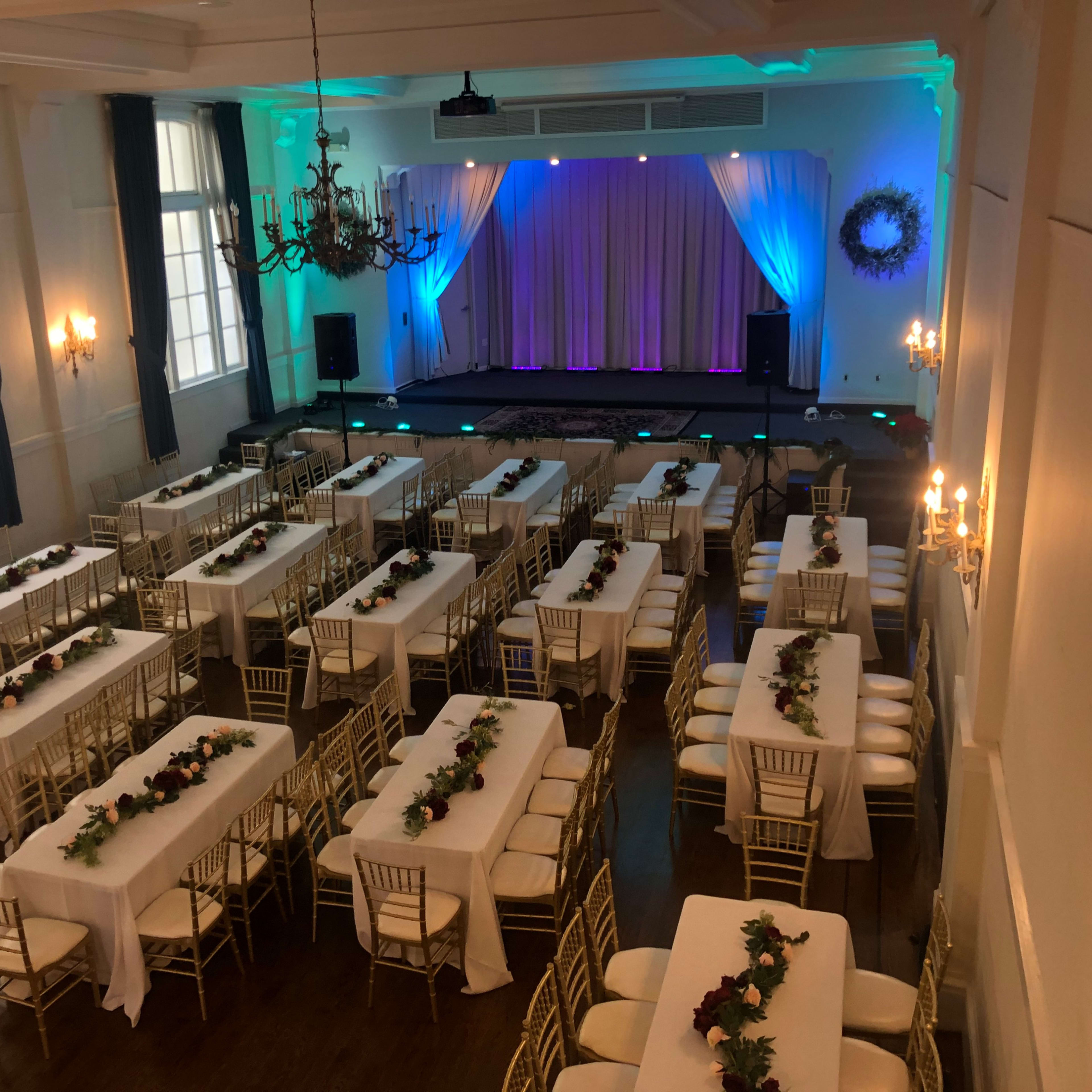 The room features rows of tables with white tablecloths and floral centerpieces, set up for an event underneath chandeliers and colored lights.