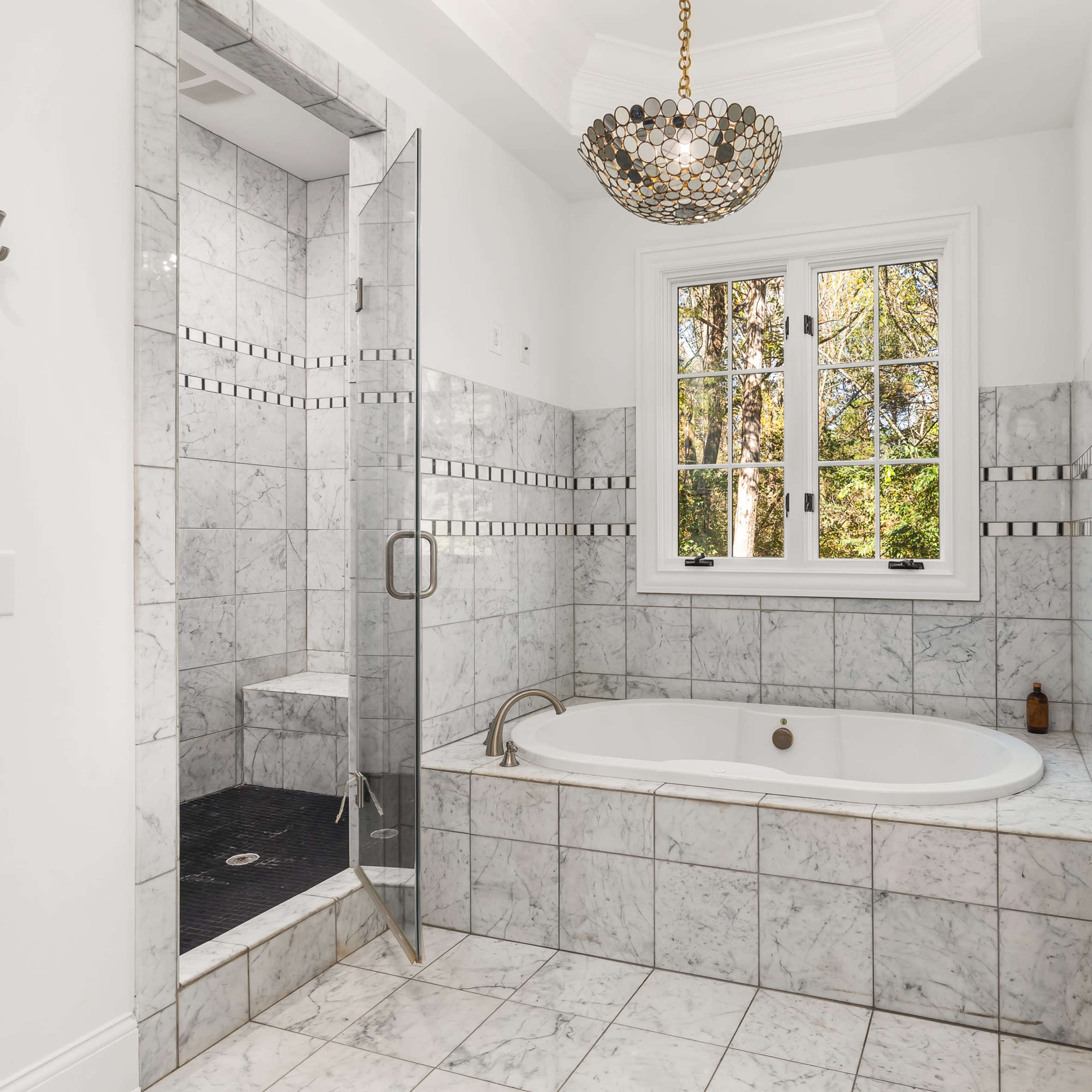 A contemporary bathroom featuring a marble tub, a glass shower enclosure, and a large window with views of greenery.