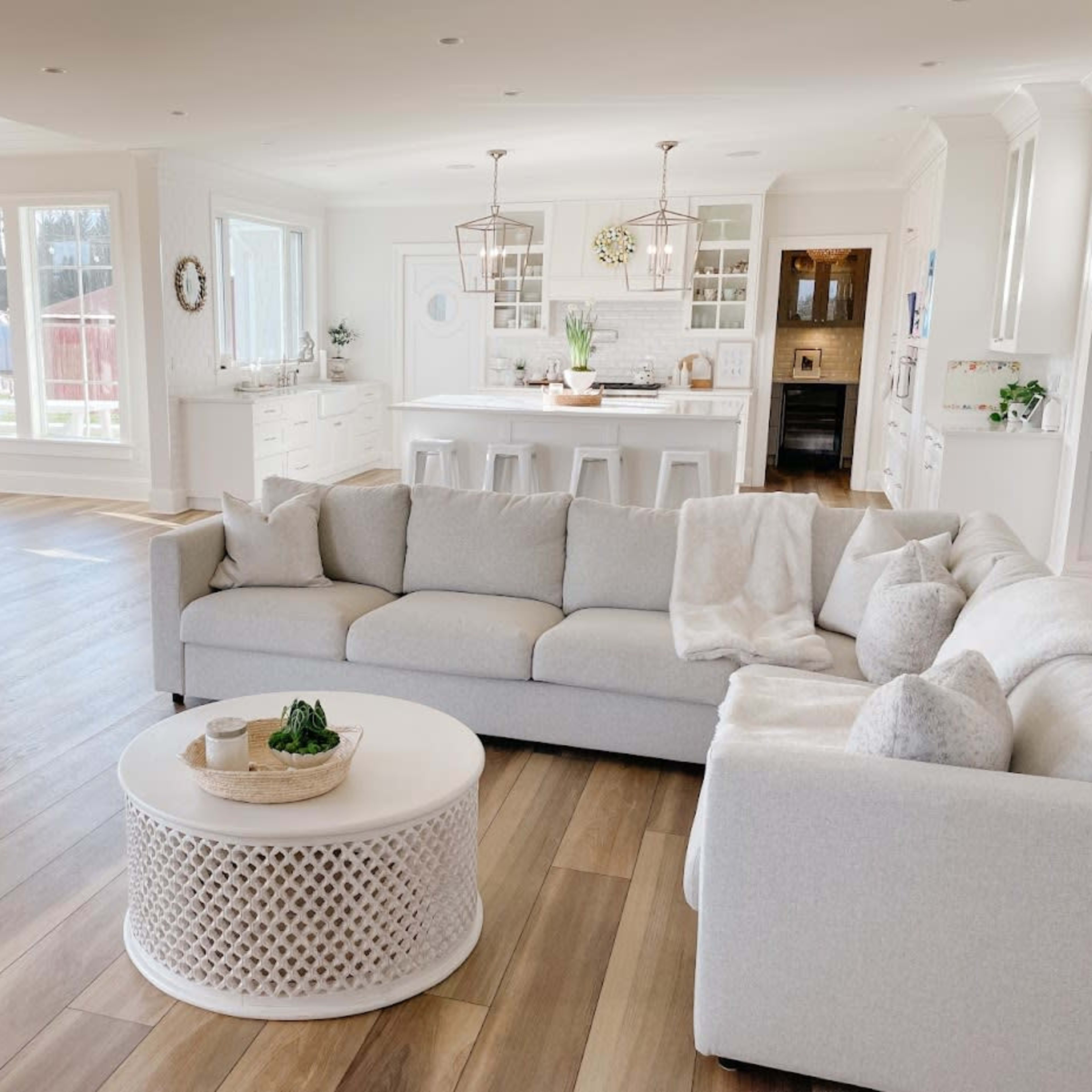 A spacious living area features a light-colored sectional sofa, a round coffee table, and large windows allowing natural light to fill the room.