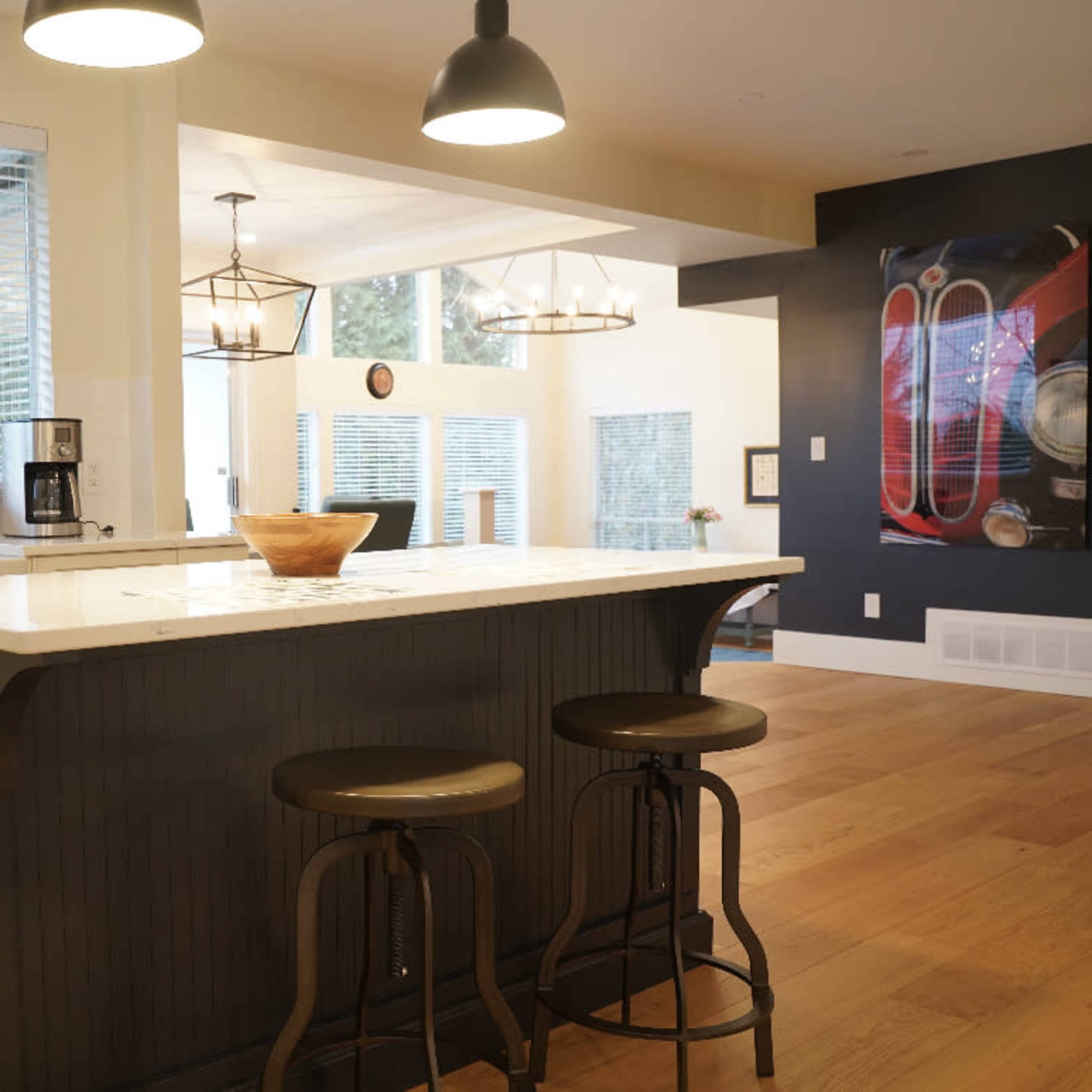 A modern kitchen with a central island, two stools, and large windows allowing natural light to enter.