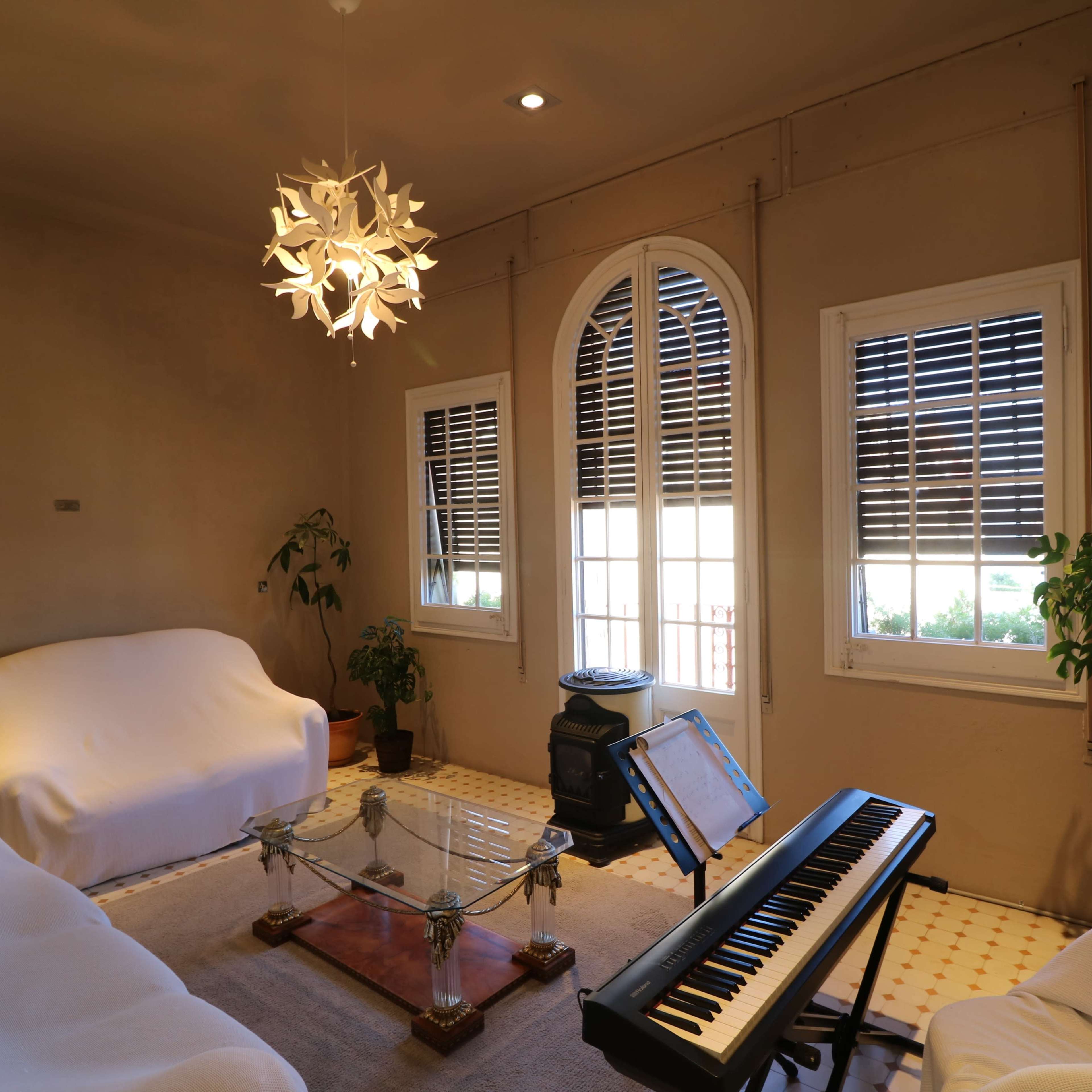 A living room features two white sofas, a glass coffee table, a piano, and large windows with shutters, all surrounded by indoor plants.