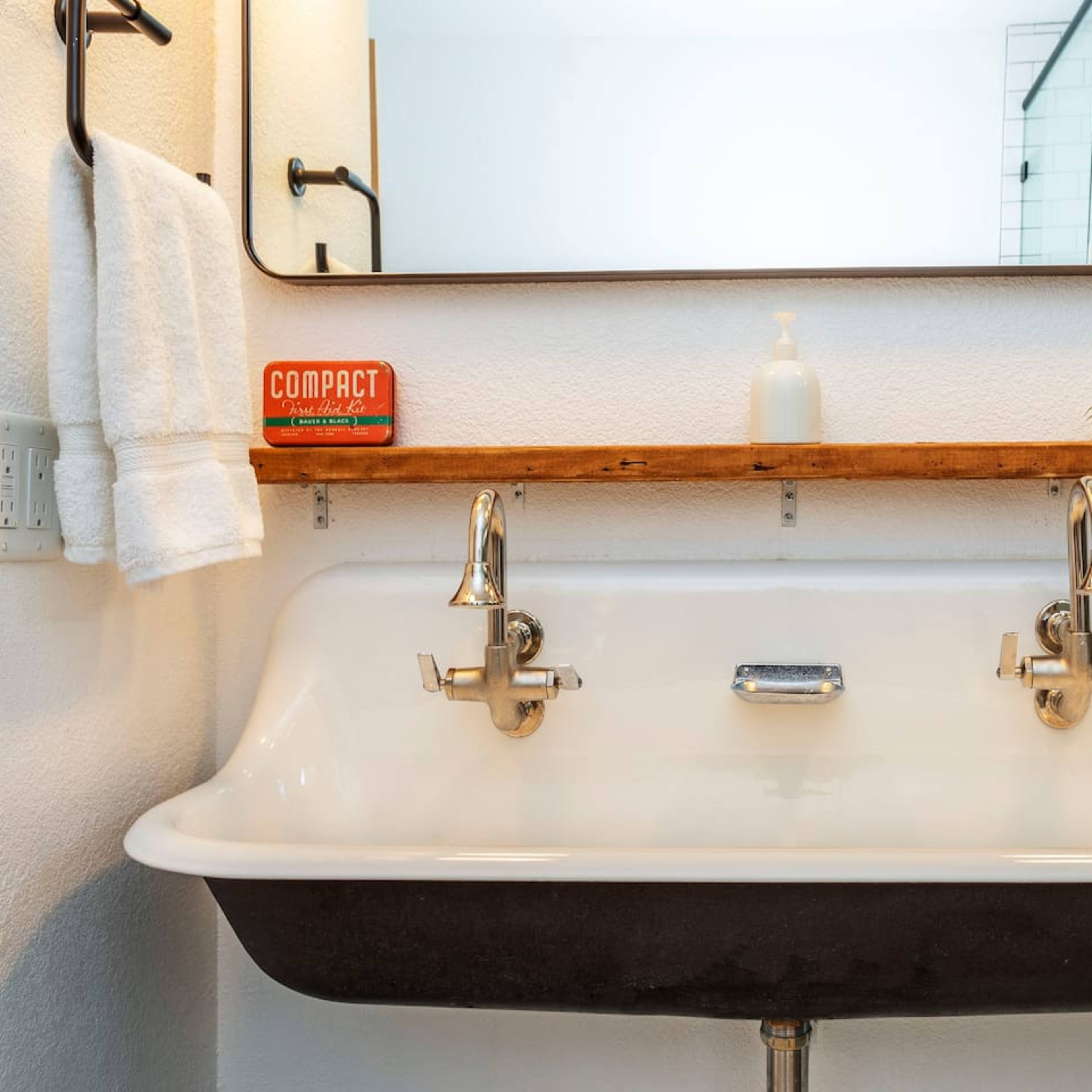 A double sink with a black basin, silver faucets, and a wooden shelf above it, complemented by a mirror and green tiled walls.