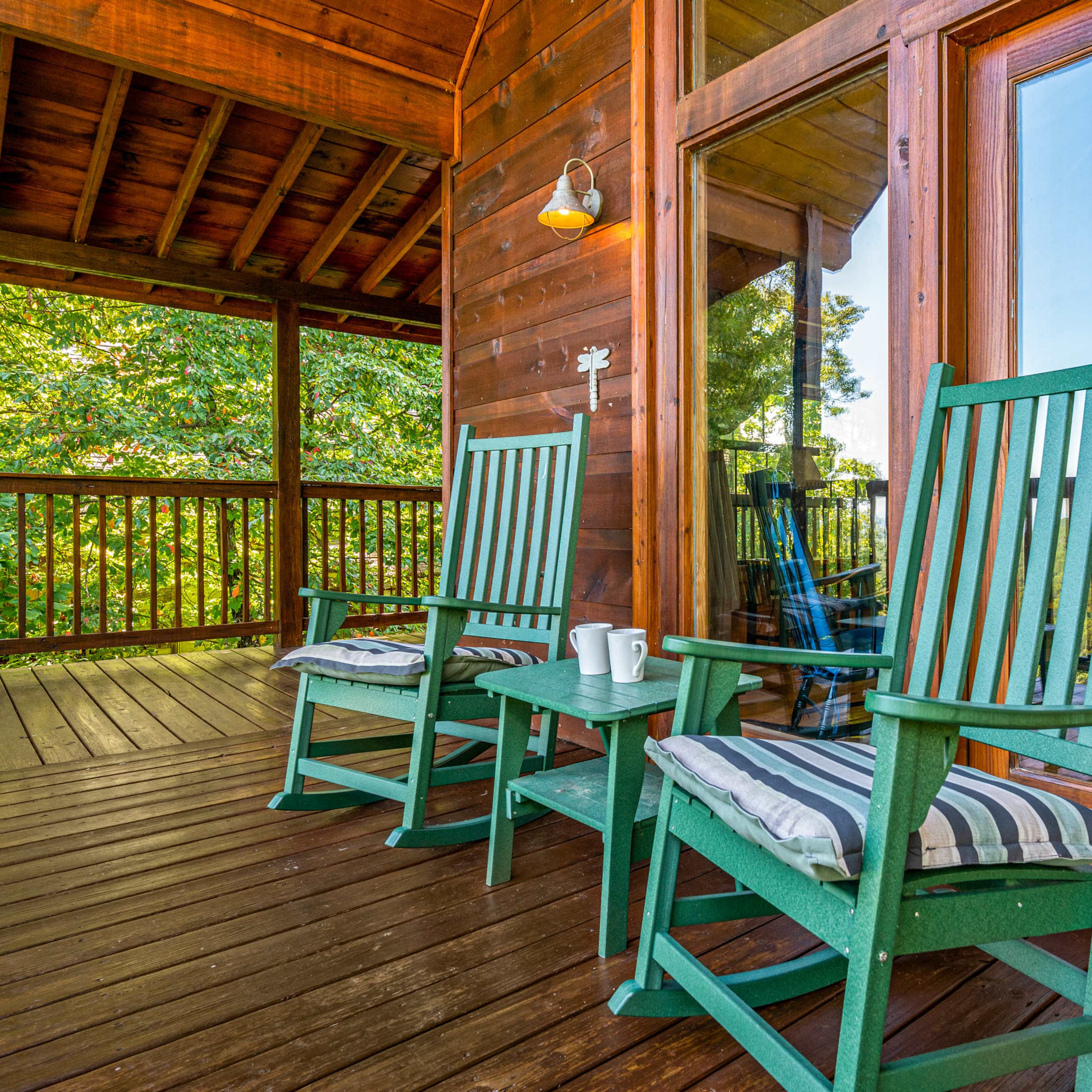 Two green rocking chairs with striped cushions sit on a wooden deck in front of large glass doors, surrounded by trees.