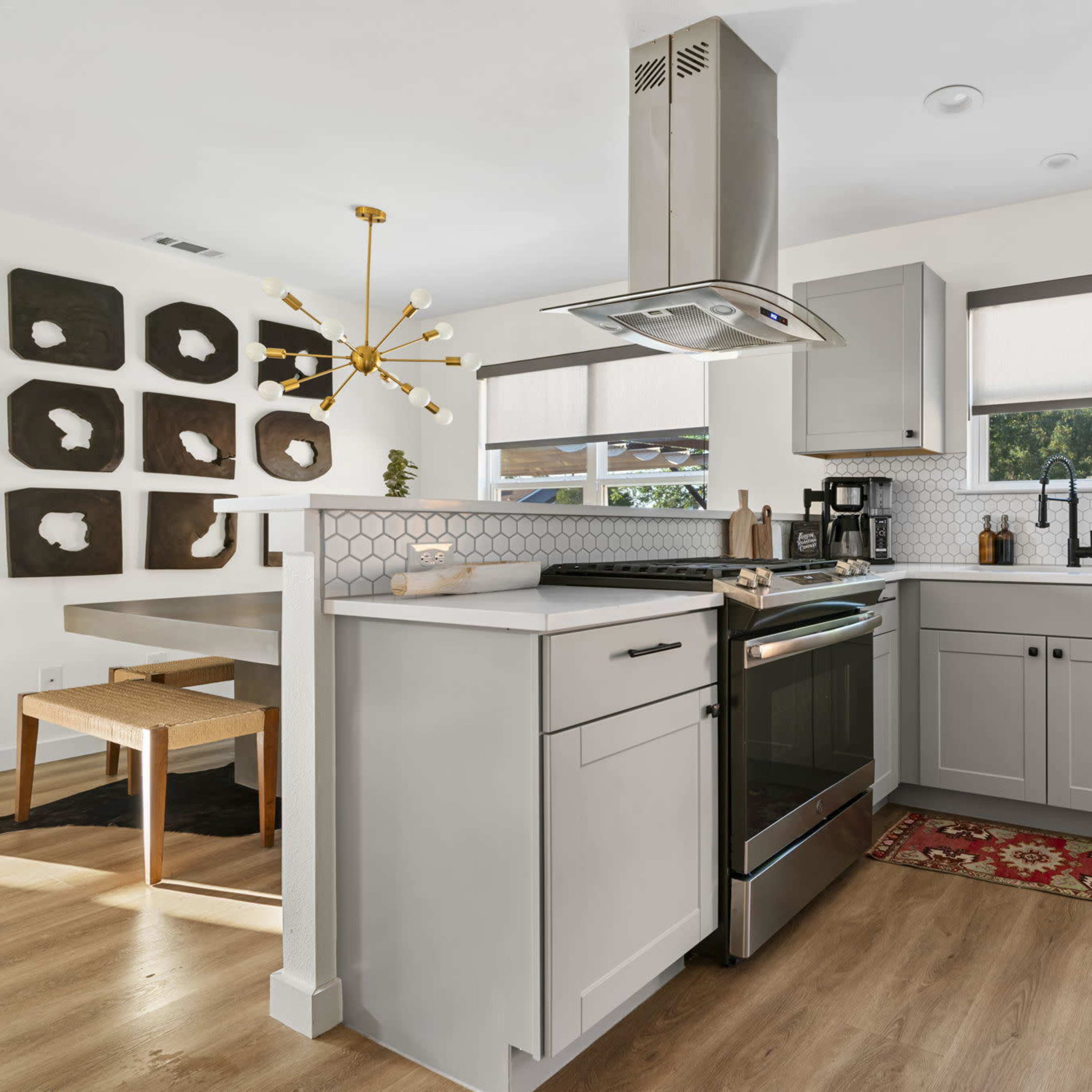 A modern kitchen features gray cabinets, a stainless steel stove, and a kitchen island with a range hood, along with a dining area that has a unique wall art display.