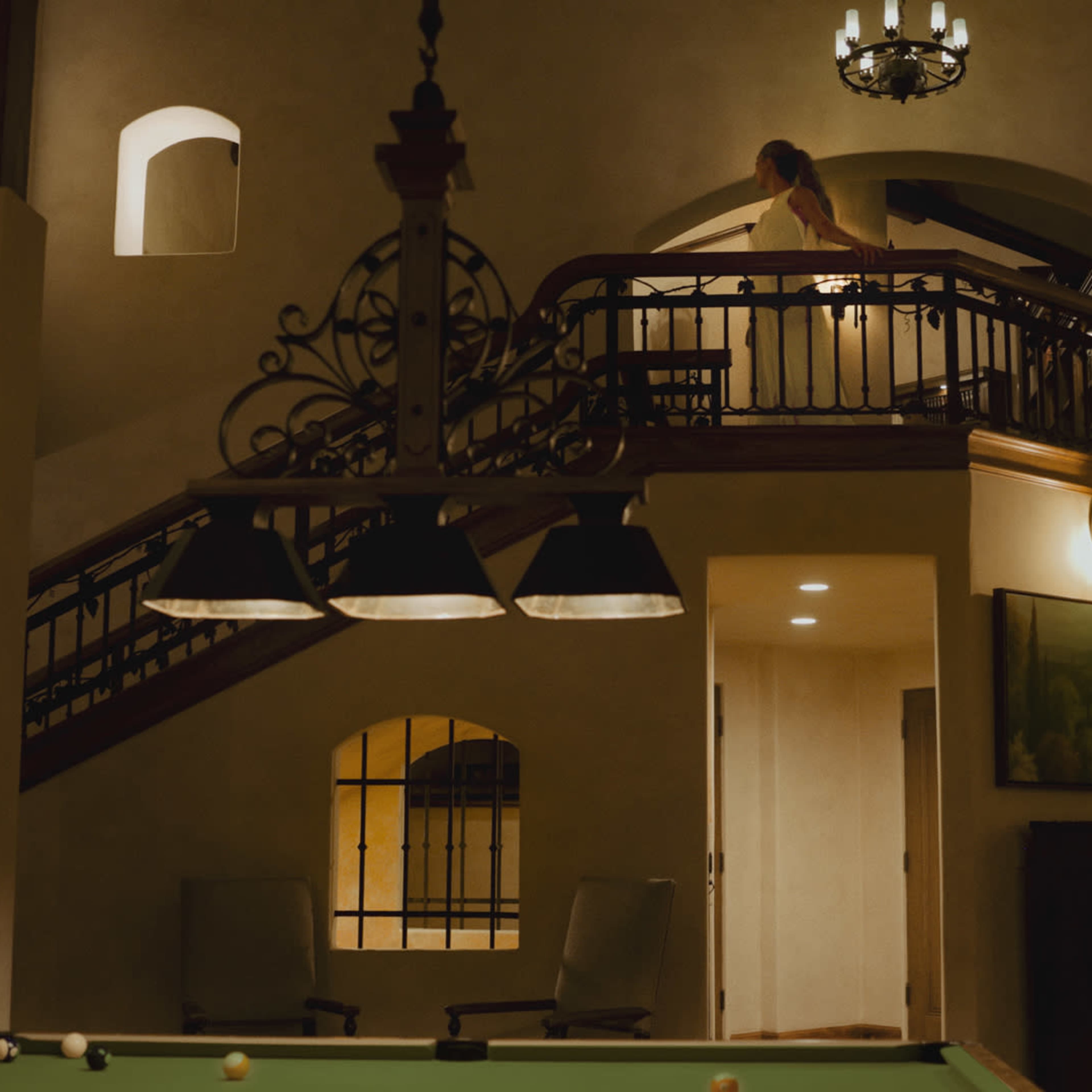 A person stands on a balcony overlooking a spacious room with a pool table beneath a chandelier.