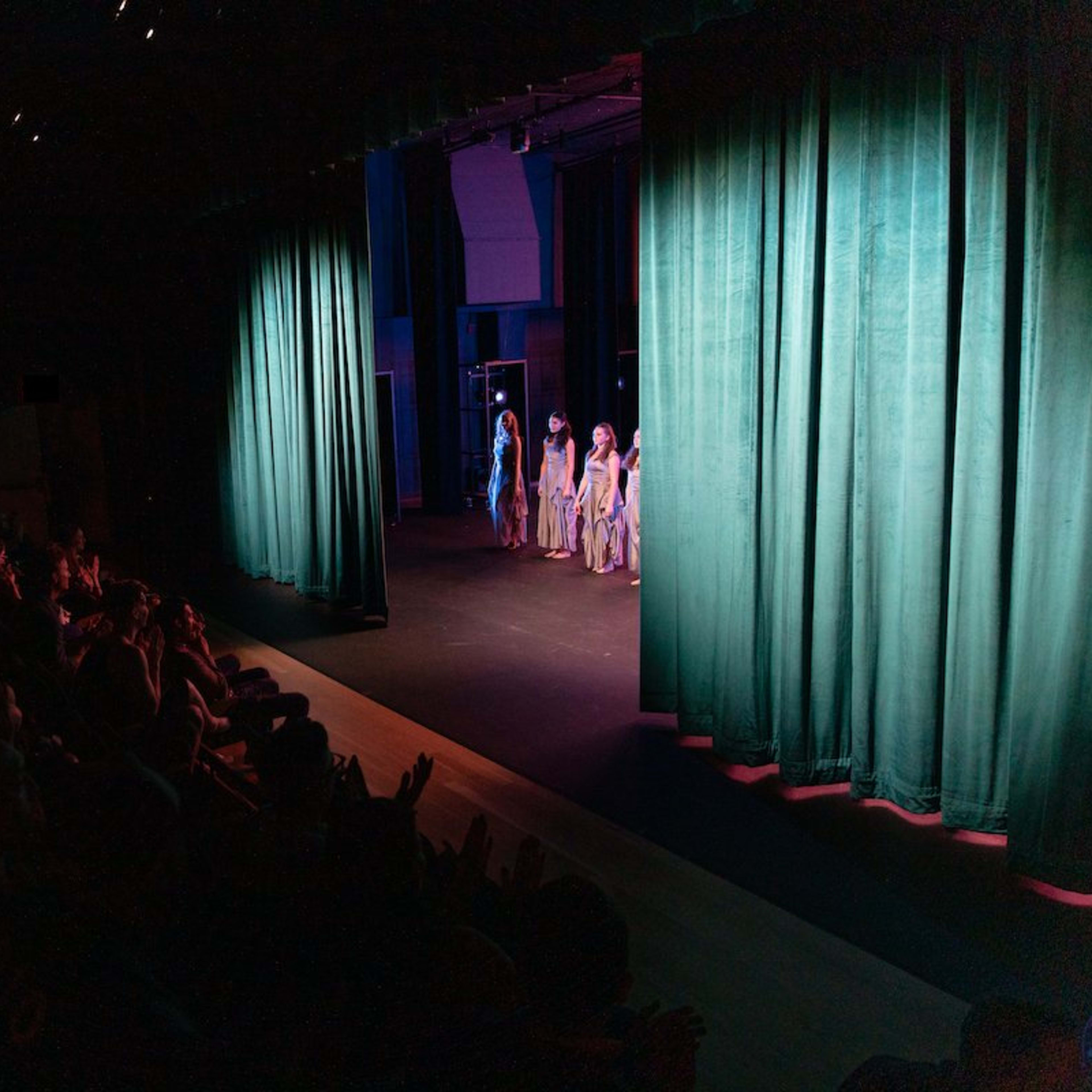 A group of performers stands on stage behind green curtains while an audience applauds from their seats.