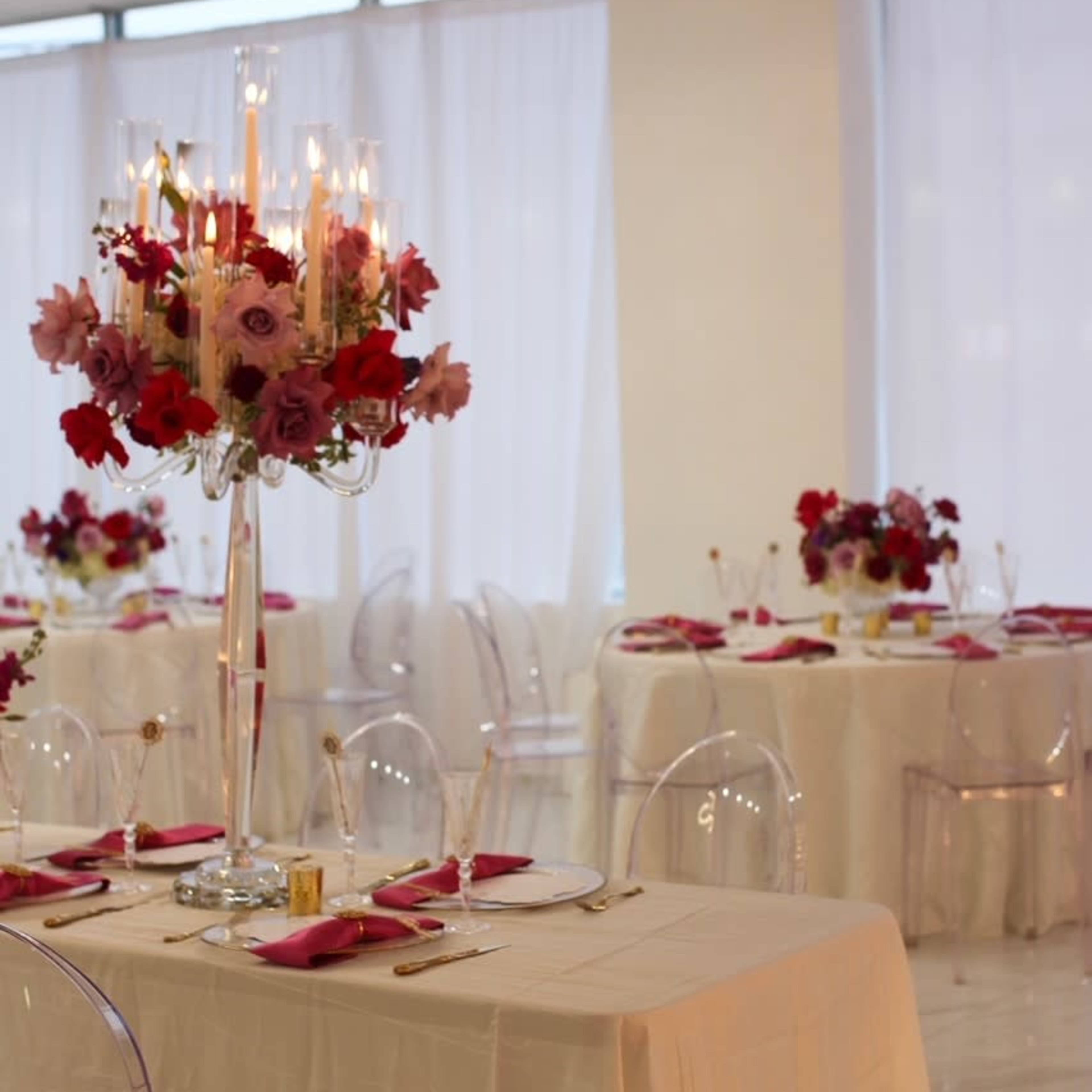 The scene features elegantly set dining tables adorned with white tablecloths, clear chairs, and elaborate floral centerpieces.