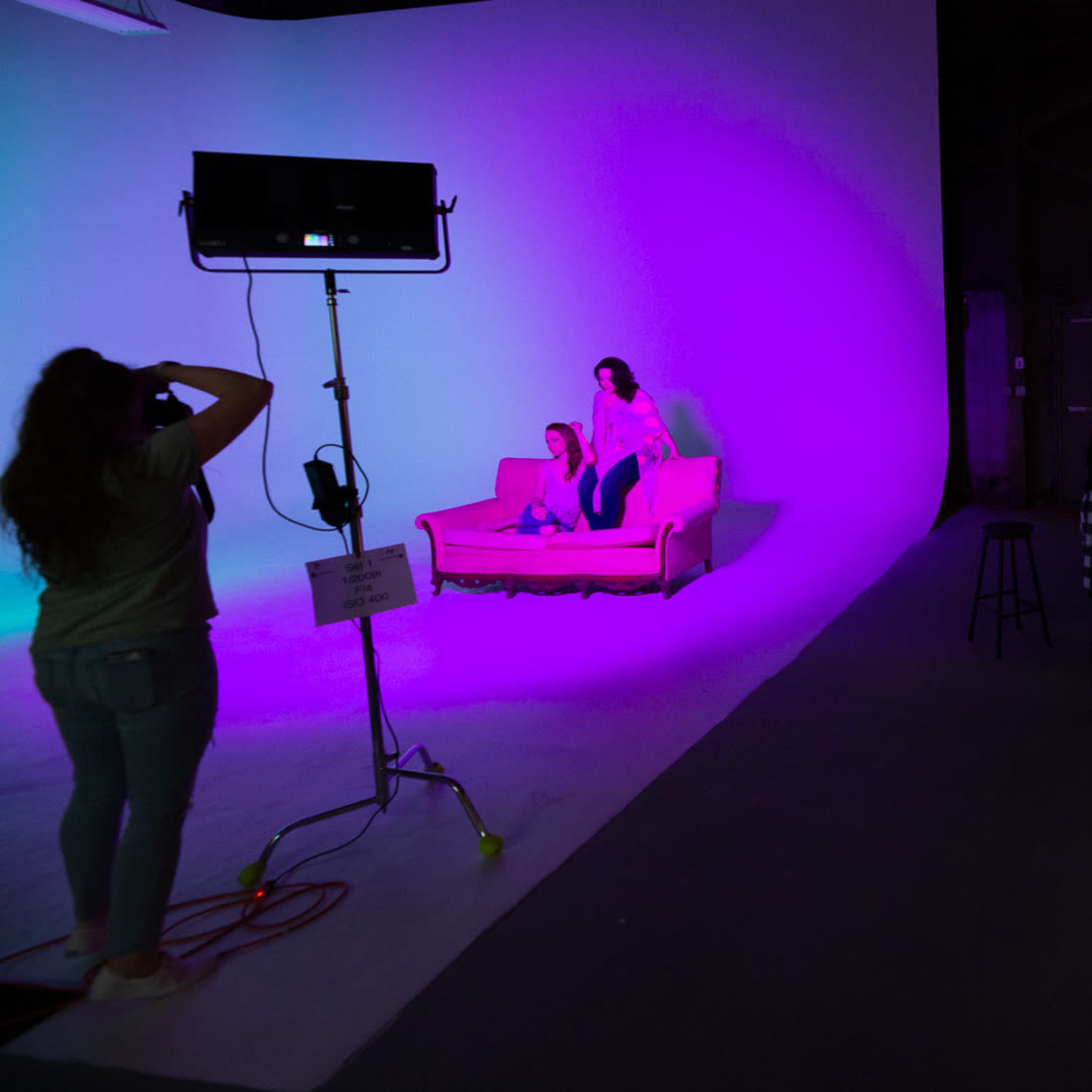 A film crew sets up a colorful studio scene with actors, a sofa, and various lighting equipment while a performer in a gold costume stands nearby.