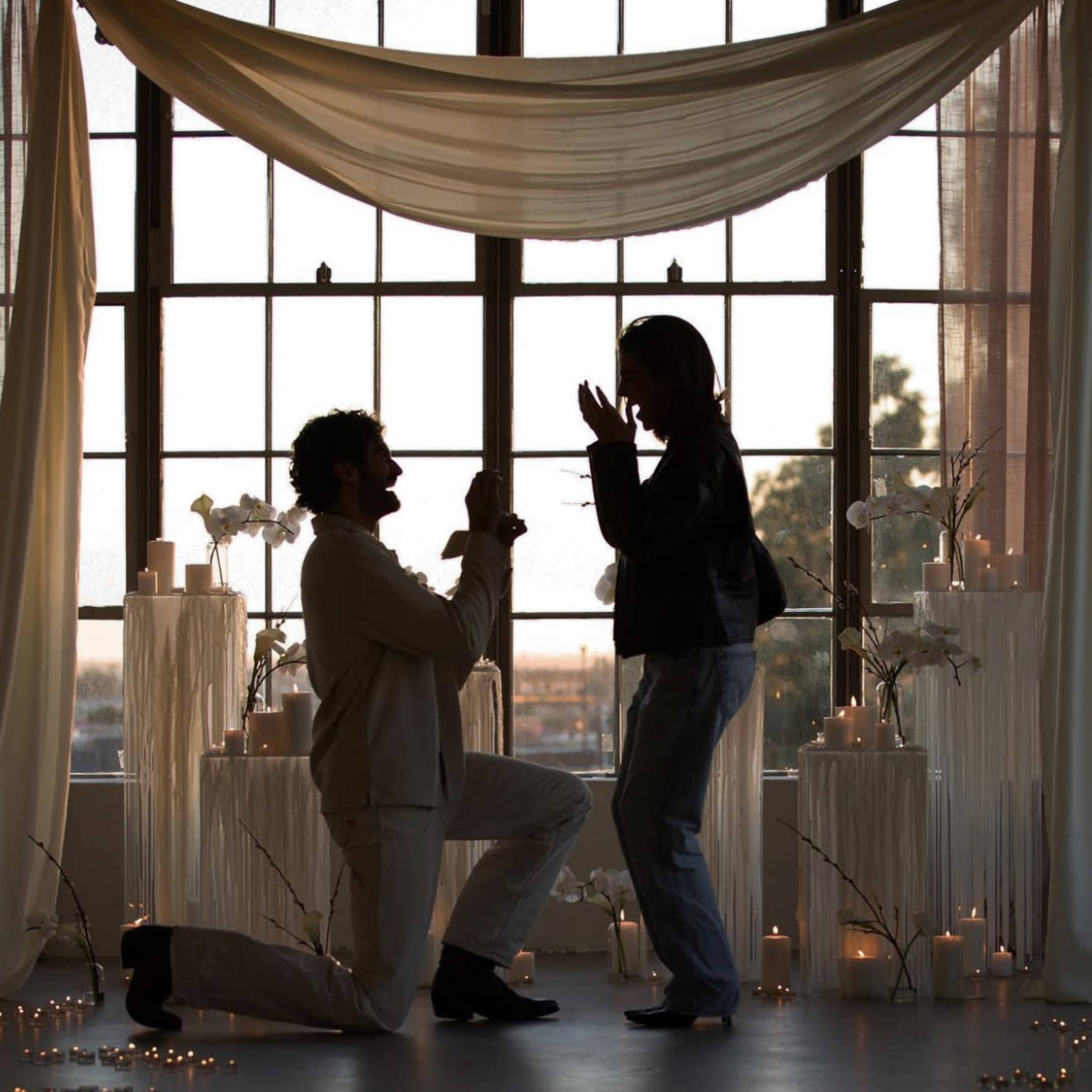 A man kneels with a ring in hand while proposing to a woman in a softly lit room decorated with candles and flowers.