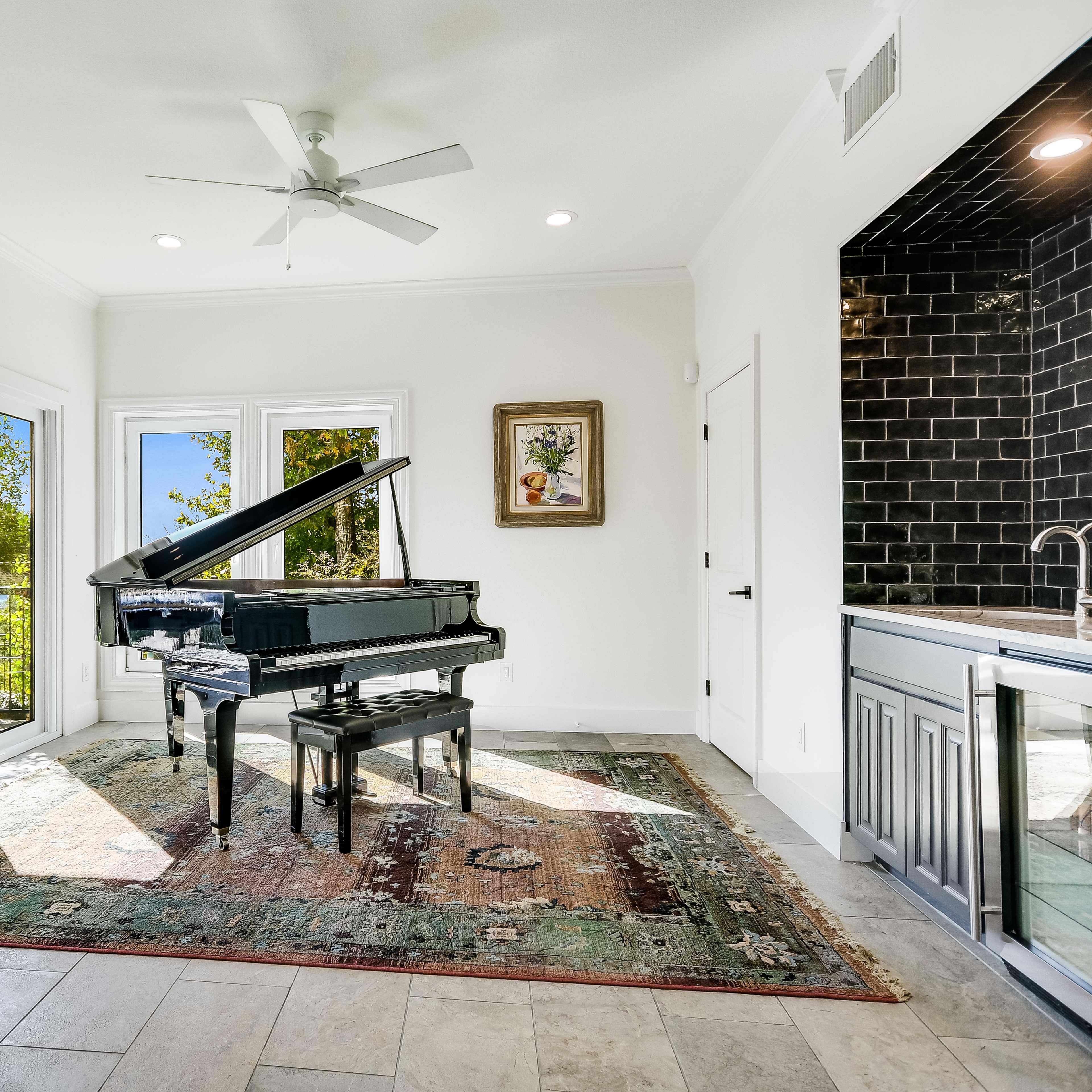 A black grand piano is situated on an ornate rug in a brightly lit room with large windows and a kitchenette in the background.
