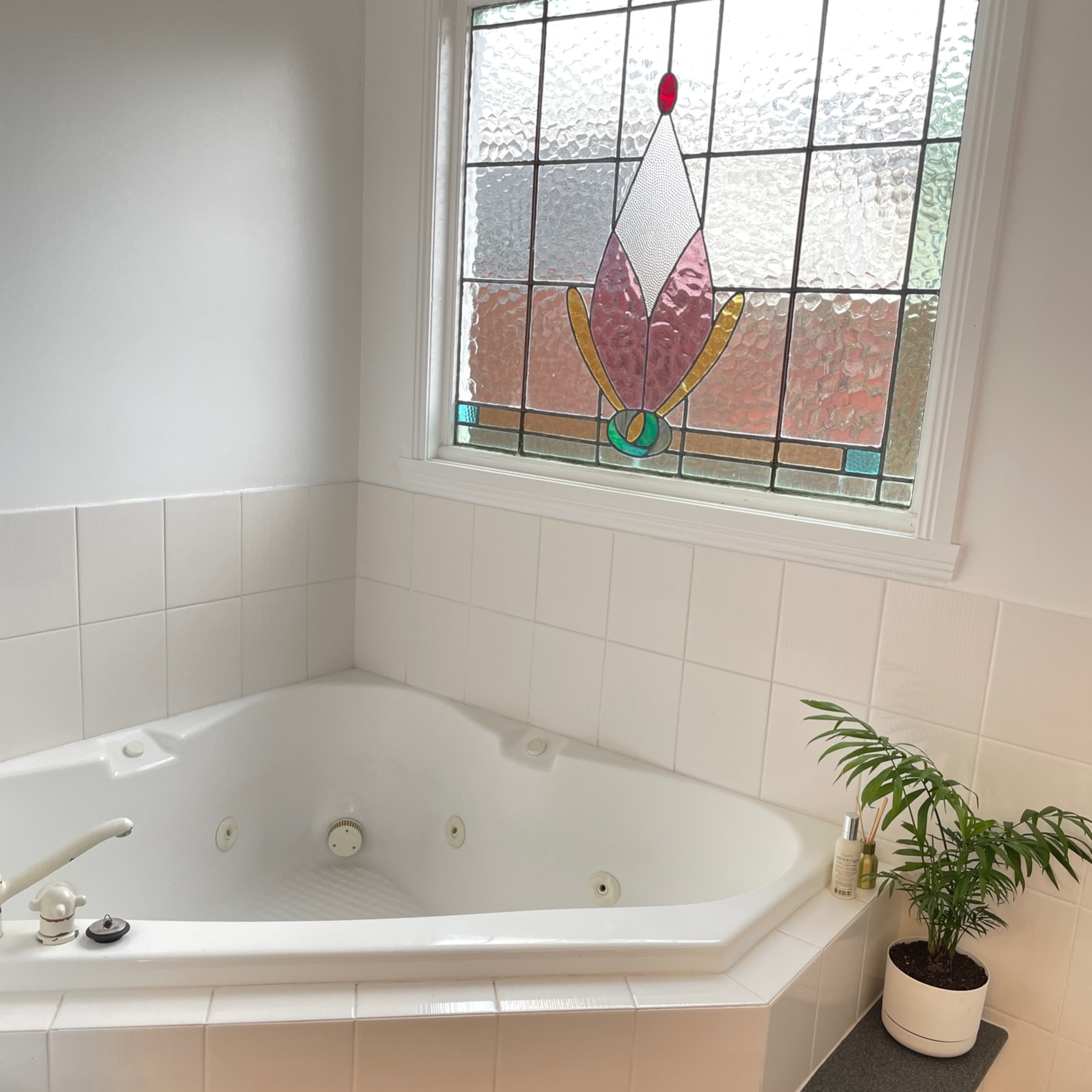 A corner jetted bathtub next to a window featuring a stained glass design, with a potted plant on a countertop nearby.