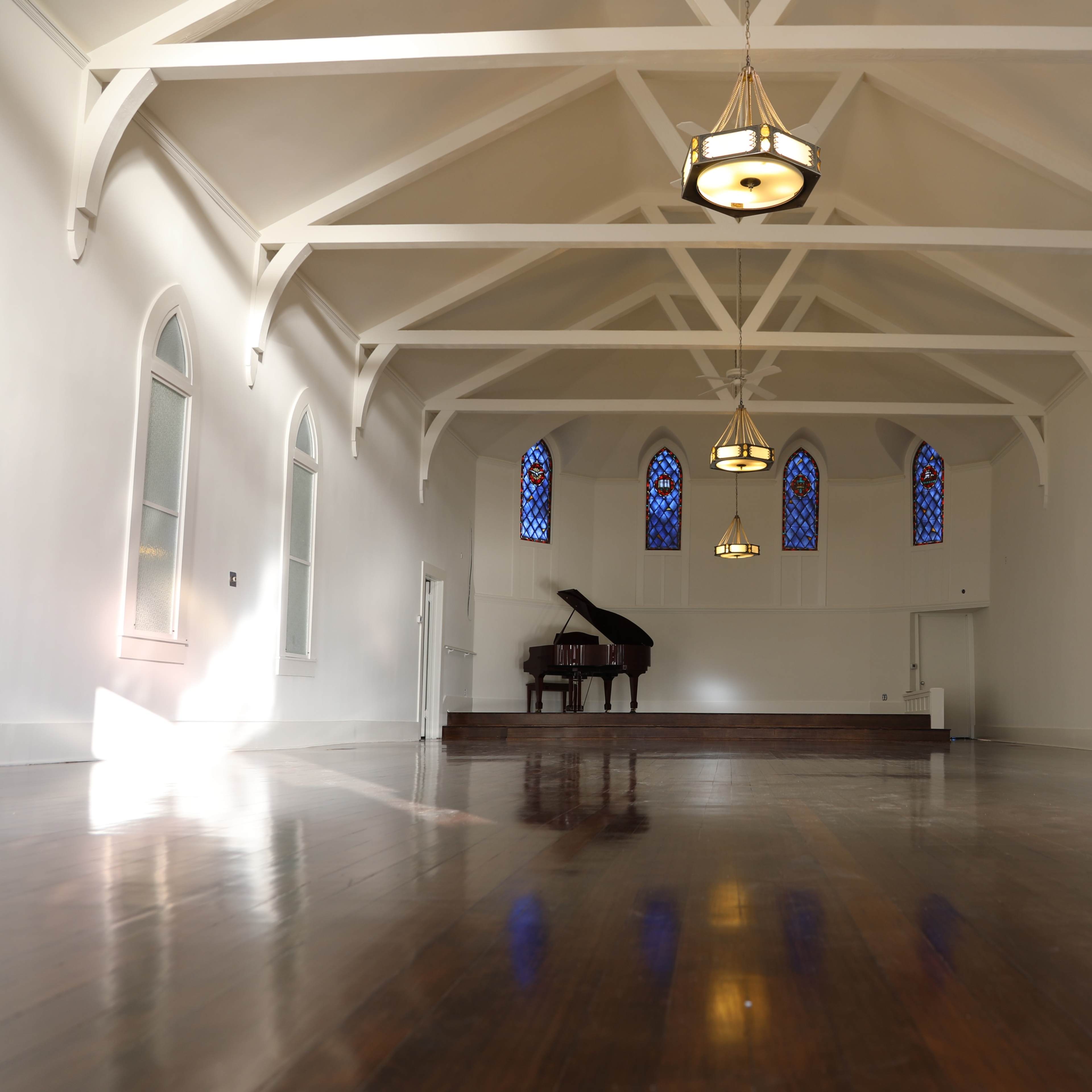 A spacious, well-lit room with wooden floors, large windows, and a grand piano on a raised platform.