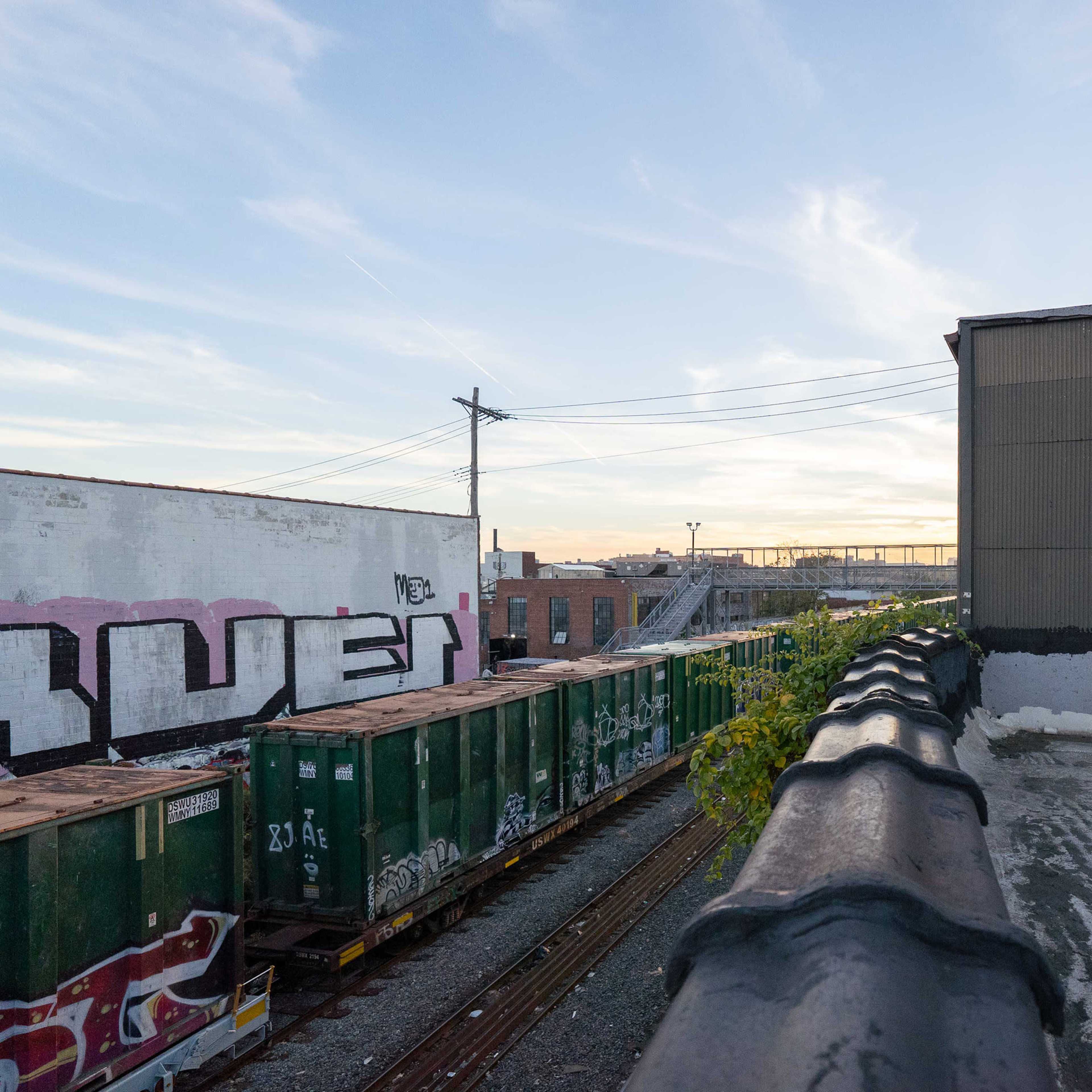 A view from a rooftop shows a line of green freight containers beside a railway, with a building and graffiti on the adjacent wall in the background.