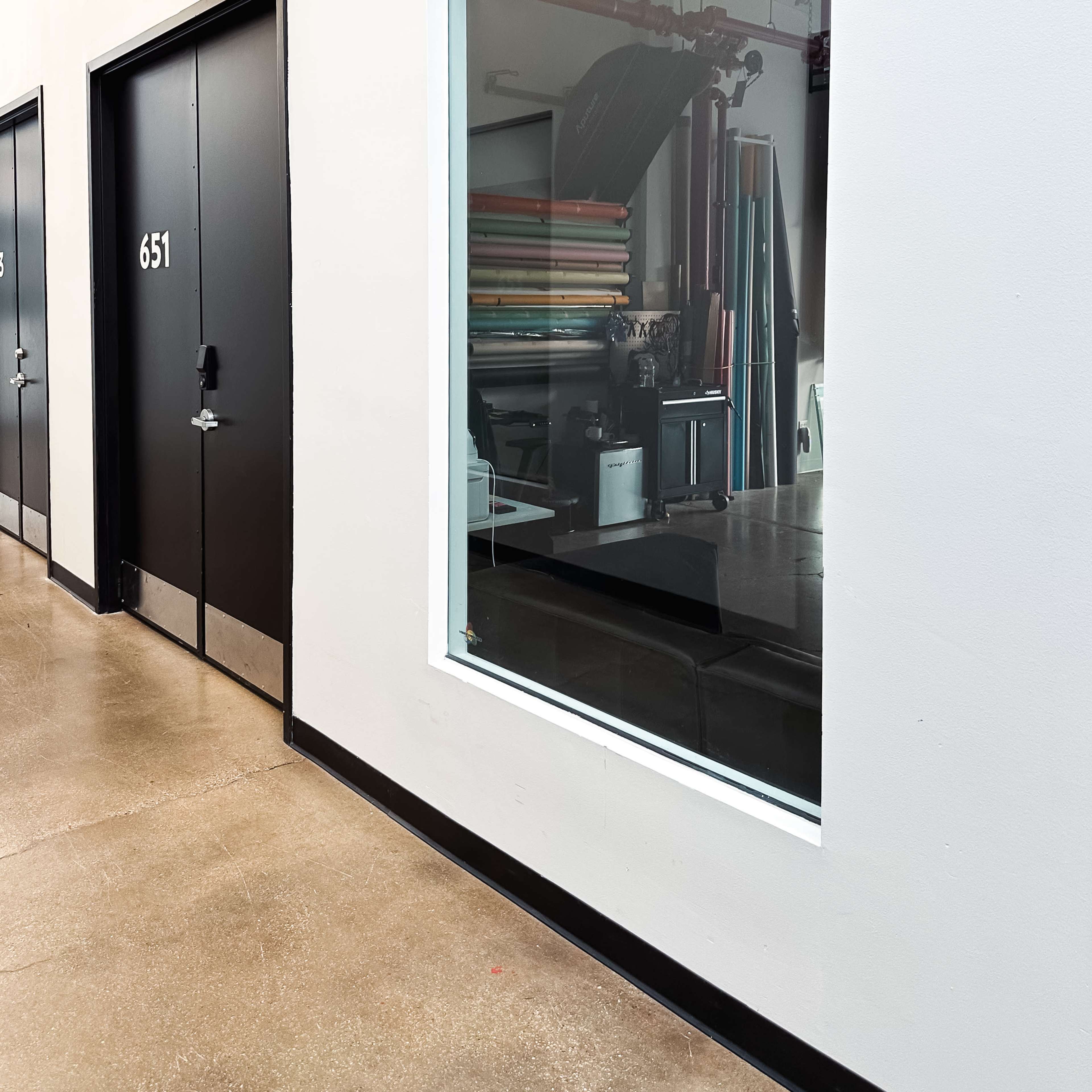 A corridor features a large window showing an interior space with various colored materials and equipment.