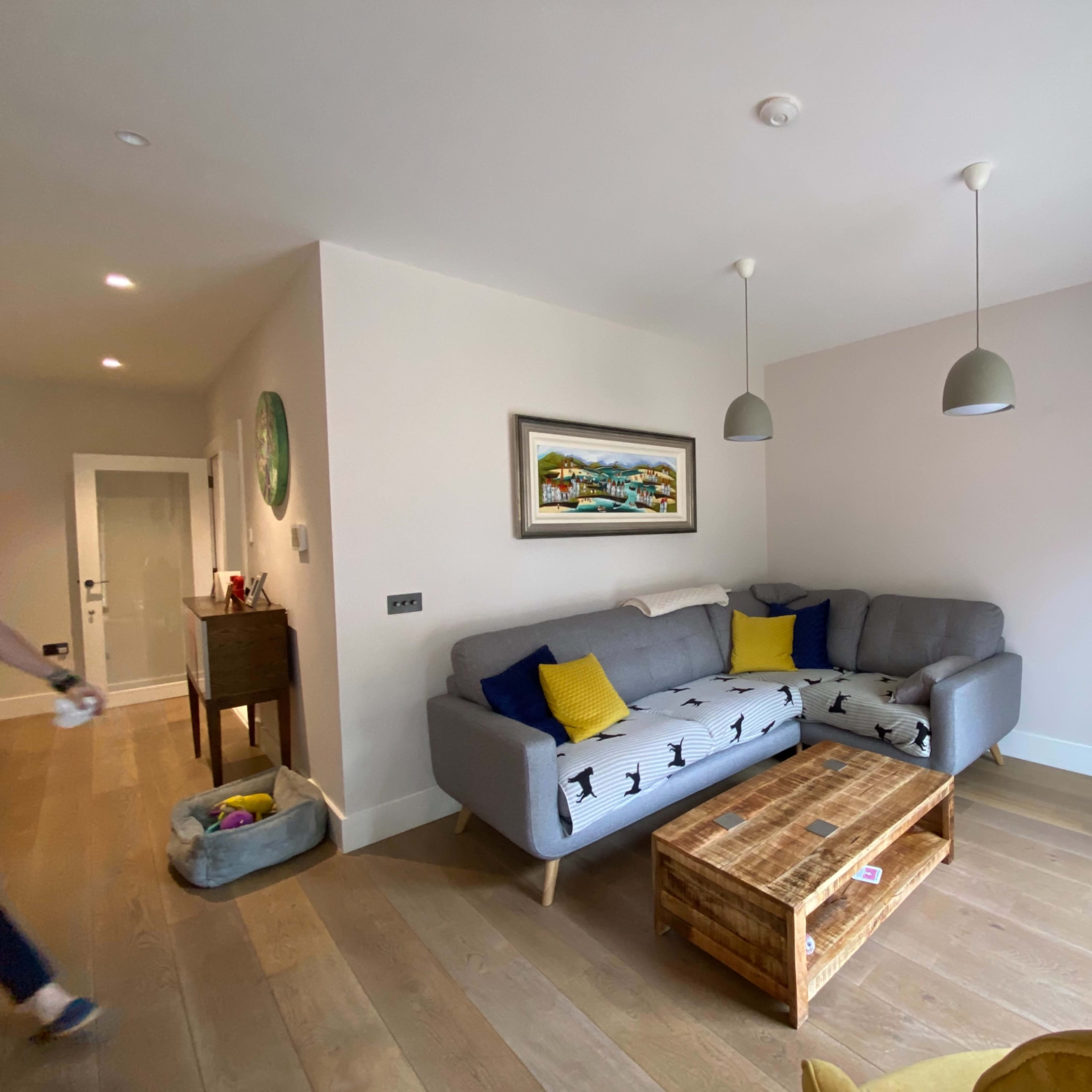 The room features a gray sofa with yellow cushions, a wooden coffee table, and a mounted television against a light-colored wall.