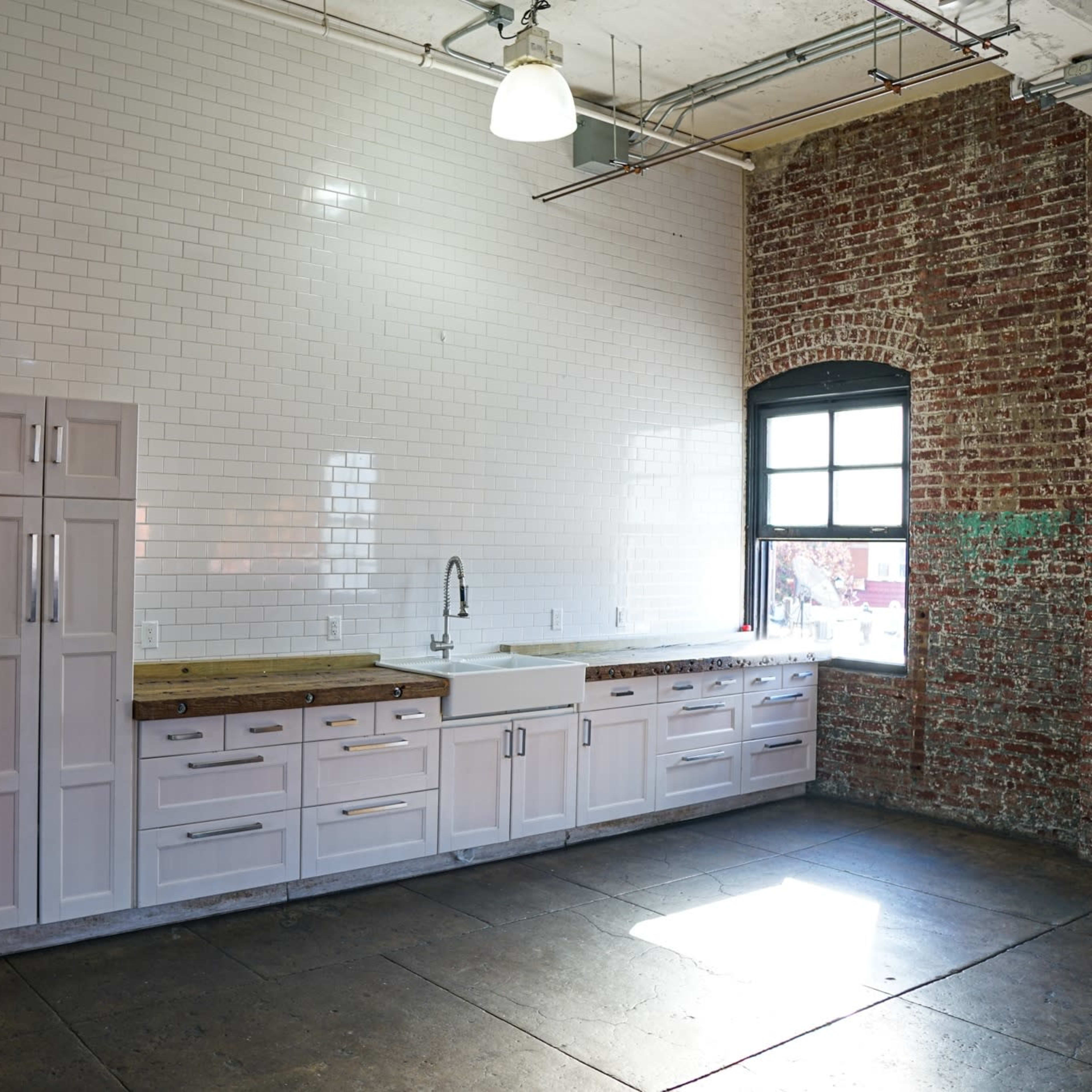 An open, industrial-style space features exposed brick walls, a kitchen area, and large mirrors along one side.