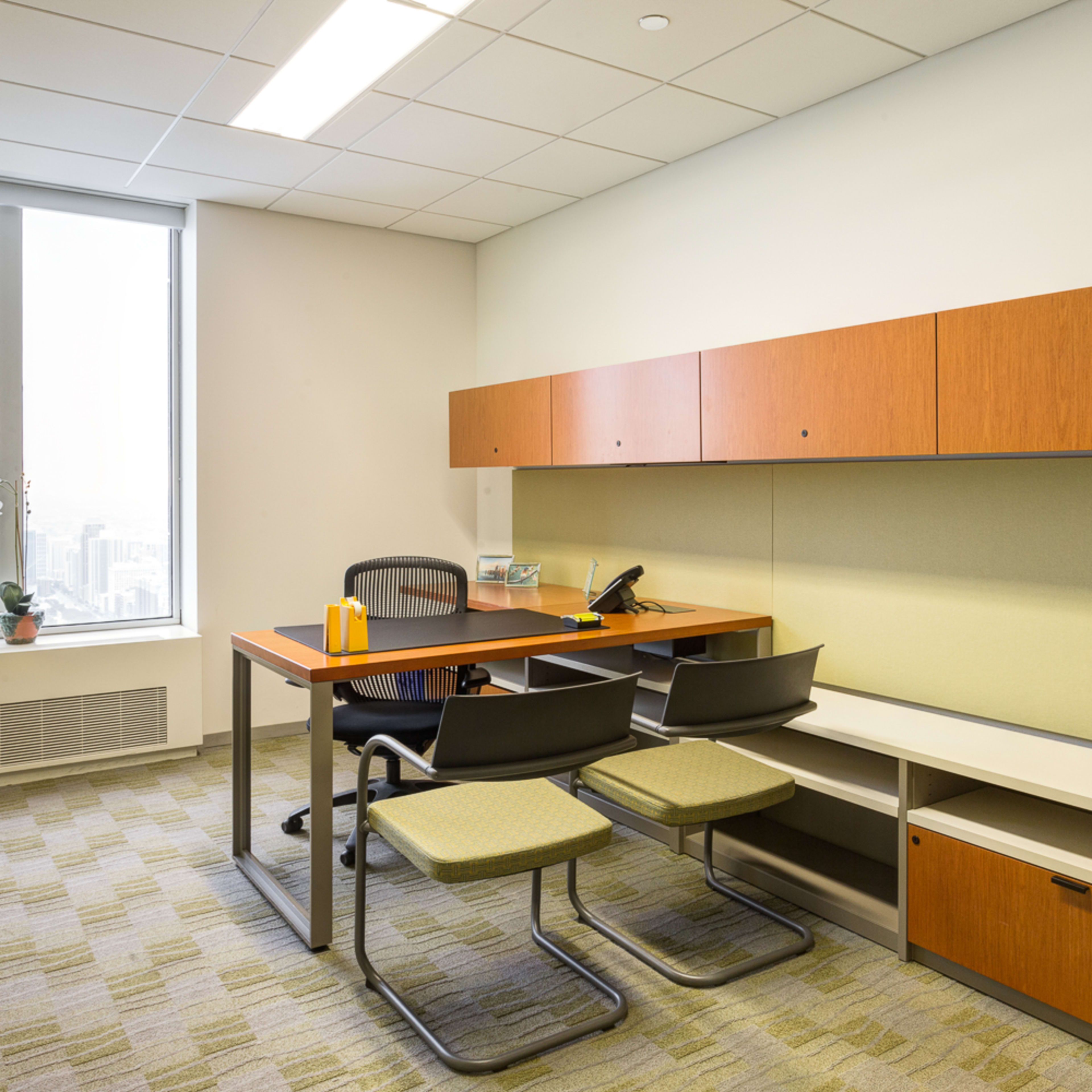 A modern office featuring a wooden desk with a chair and two additional chairs, a wall-mounted storage unit, and a large window offering a view of the city.