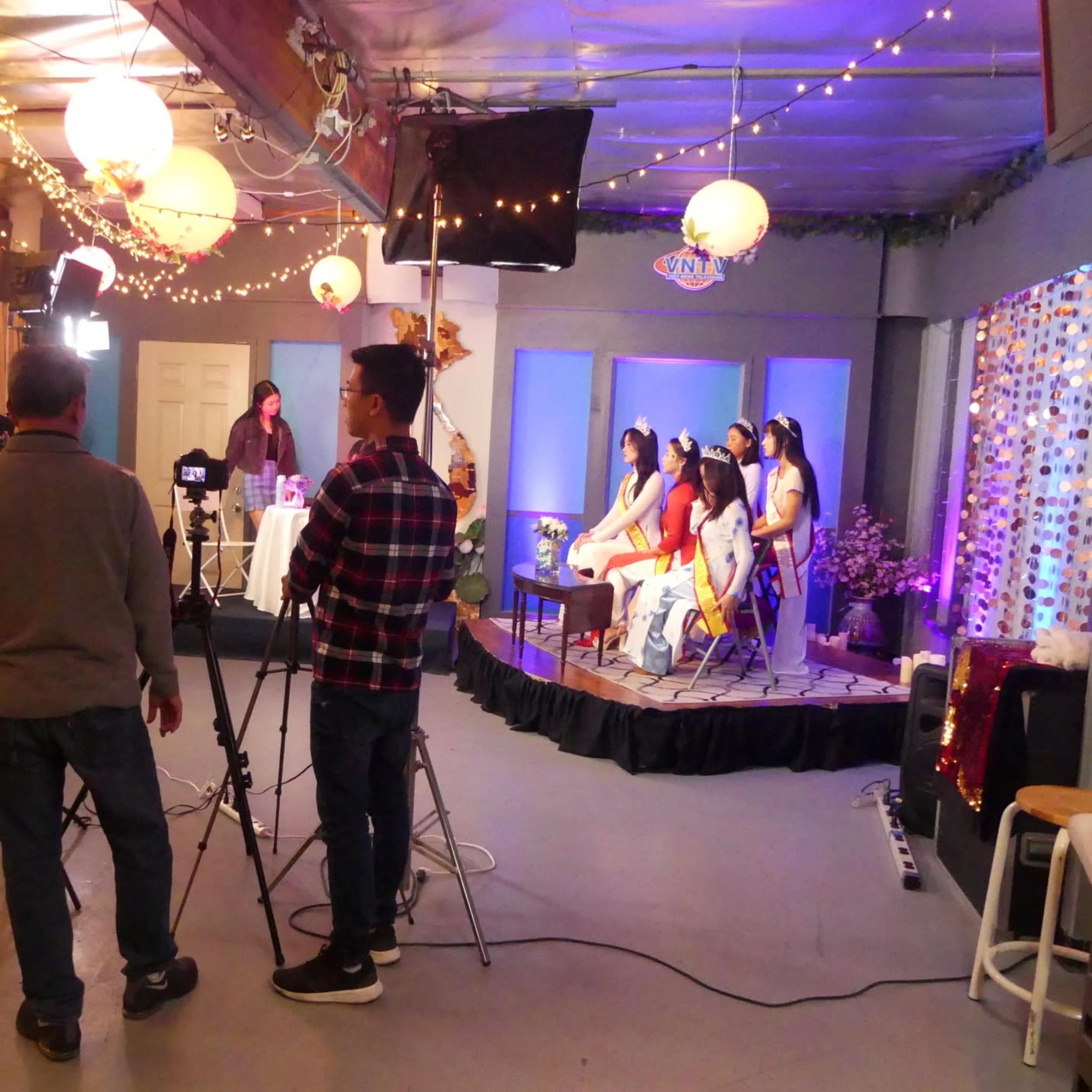 A production crew sets up lights and cameras for a staged performance featuring several seated performers in a decorated studio.