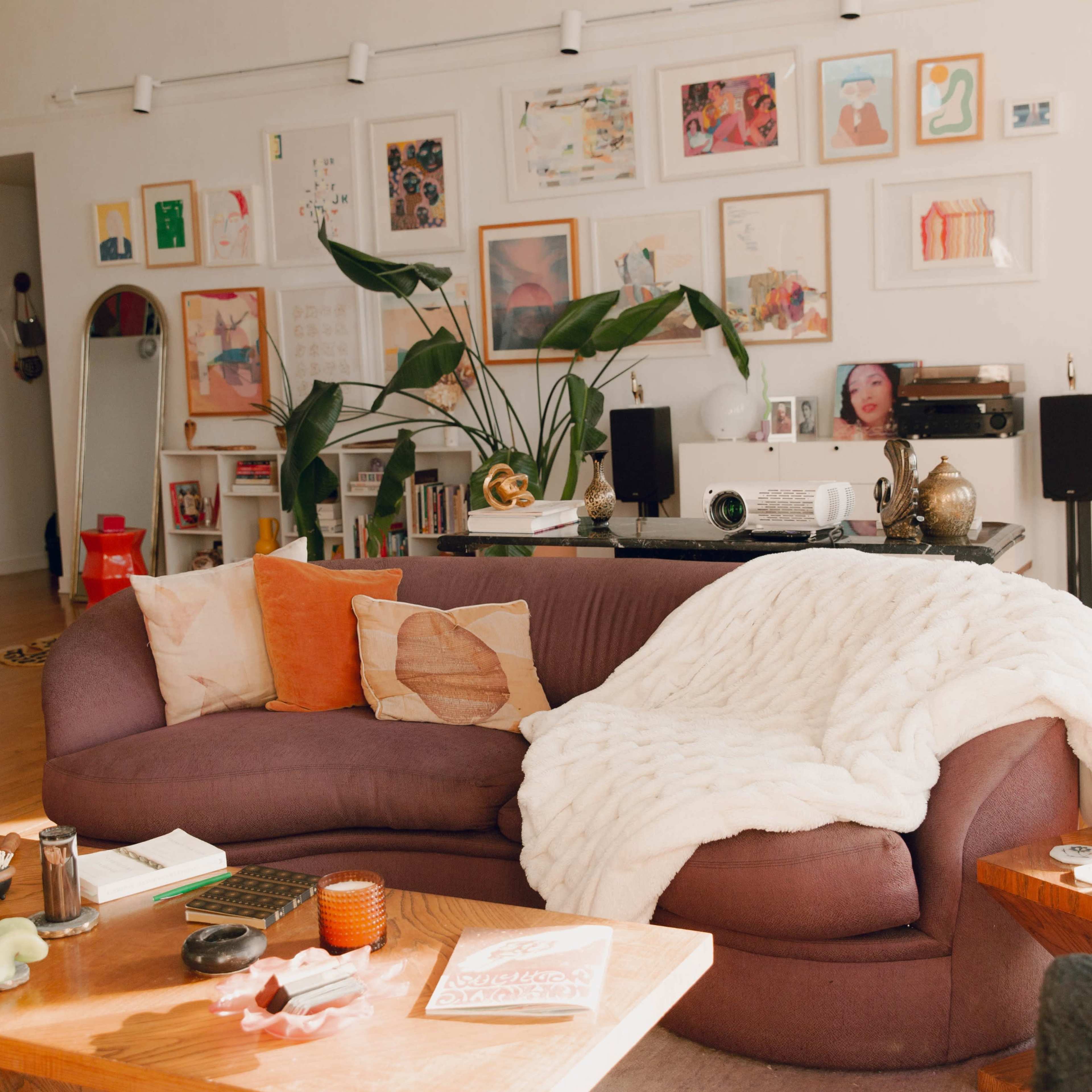 A cozy living room features a curved sofa with a throw blanket, surrounded by various art pieces on the walls and decor items on wooden tables.