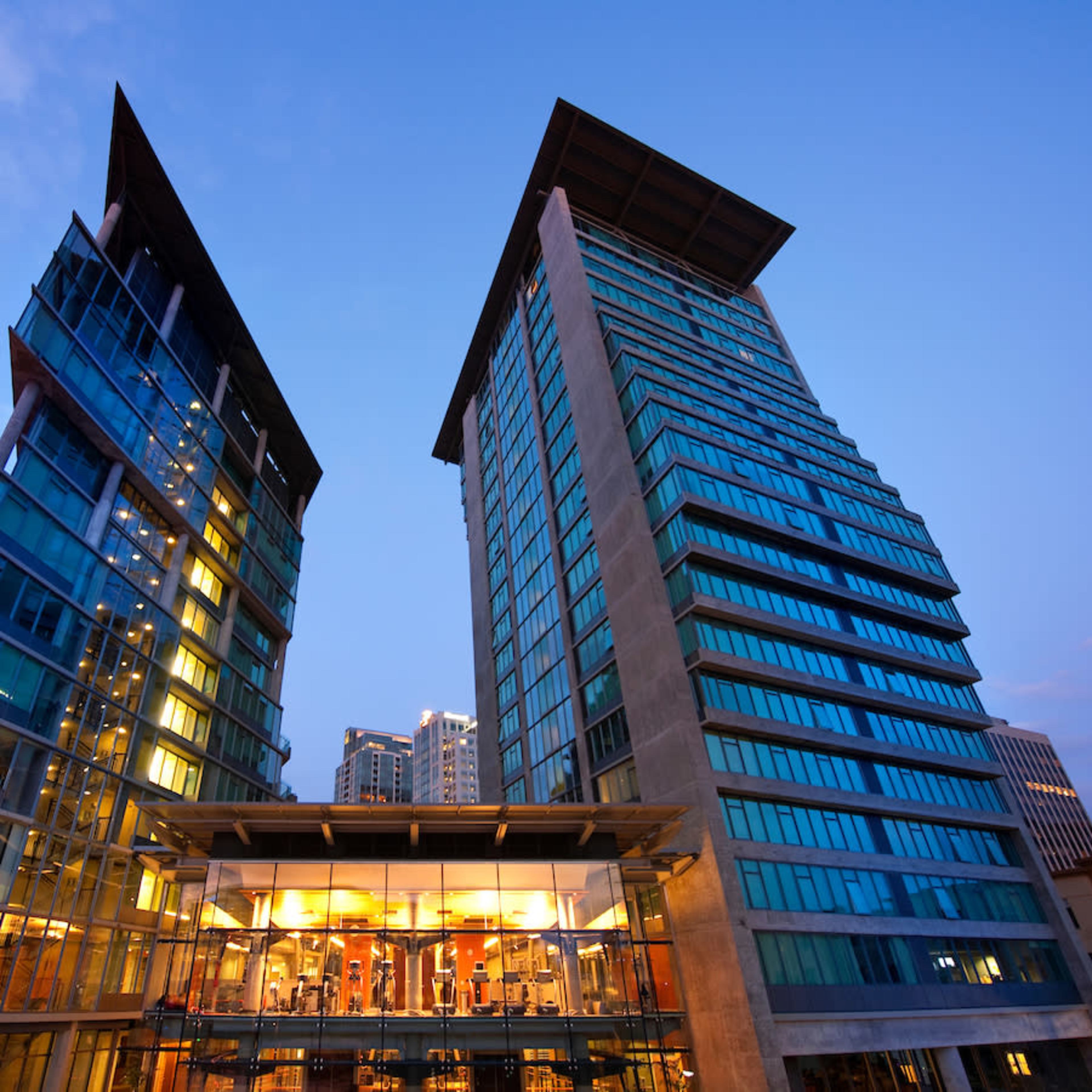 Two modern high-rise buildings with glass facades and a connected lower level illuminated at dusk.