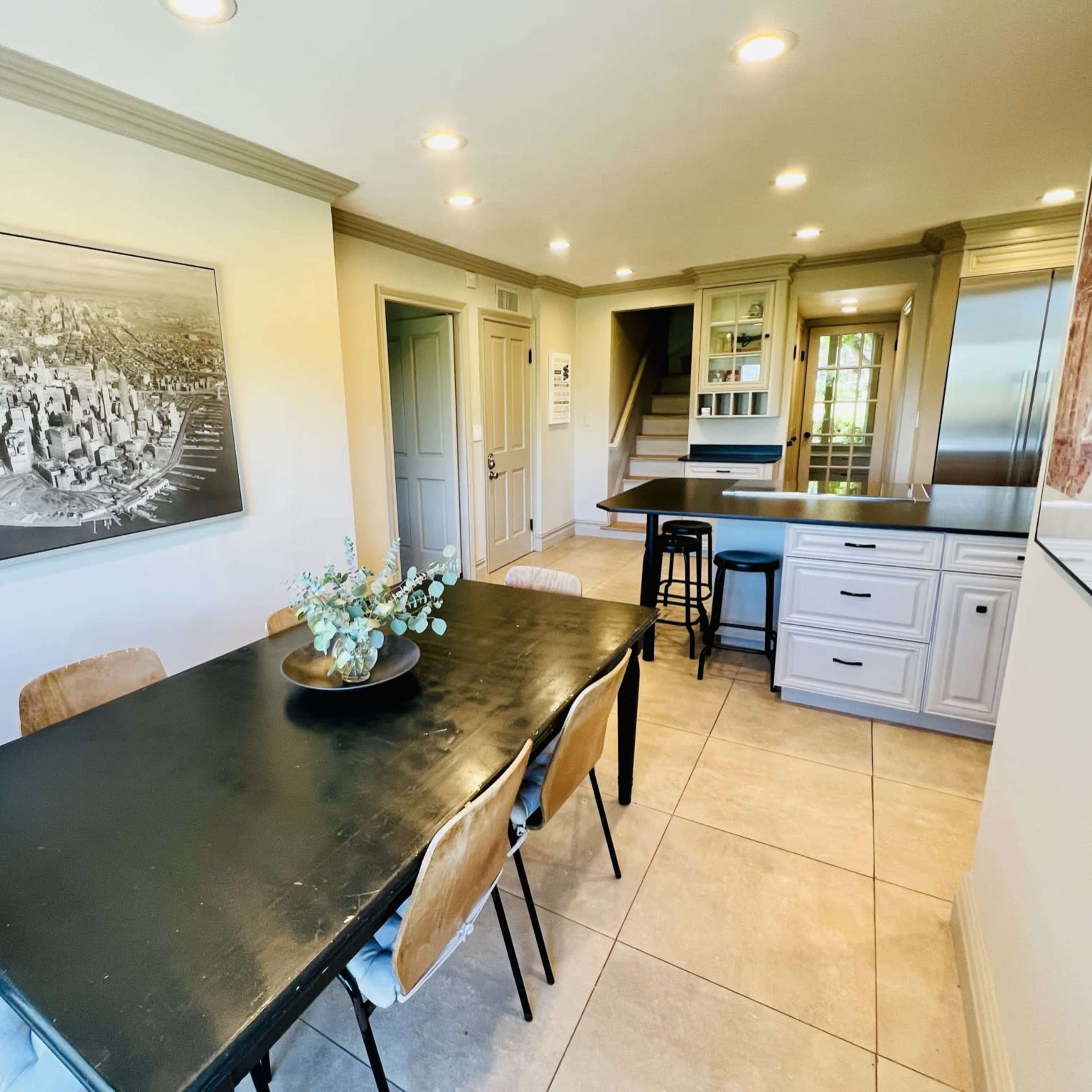 A kitchen and dining area features a large black table surrounded by wooden chairs, with cabinets and a countertop visible in the background.