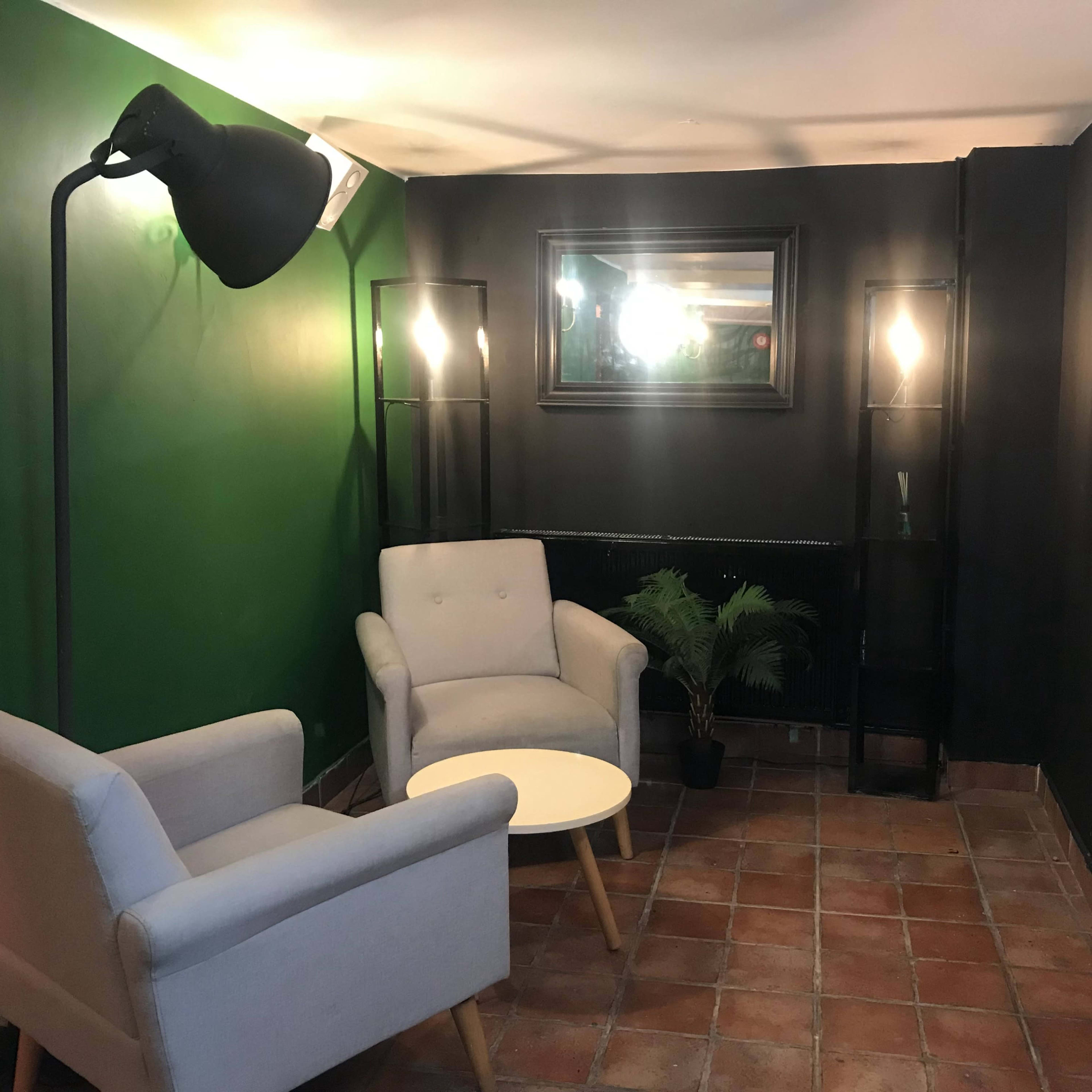 A cozy seating area with two light gray armchairs, a small circular table, and decorative floor lamps against a dark green and black wall.