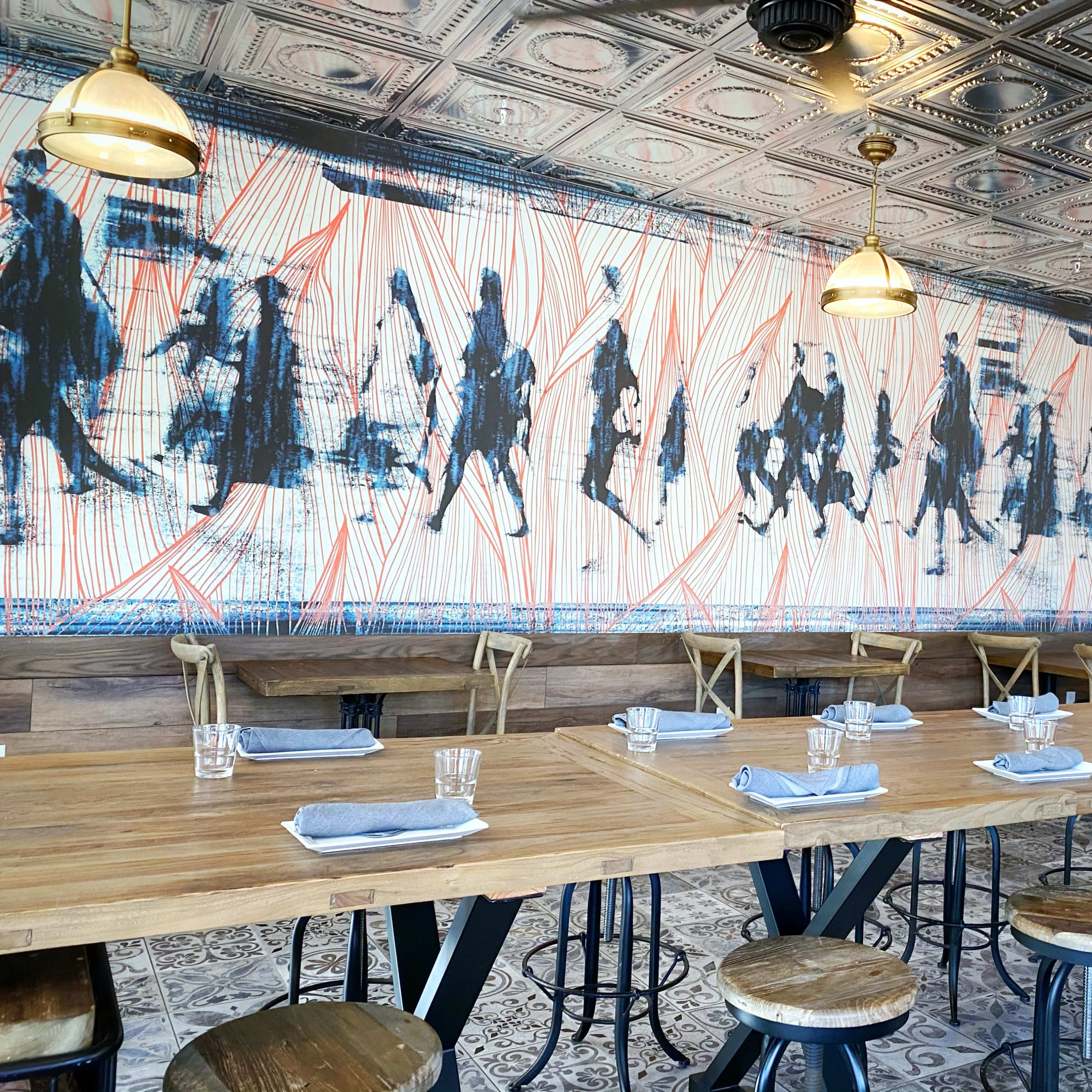 A dining area features a long wooden table set with glasses and napkins, surrounded by stools, and is adorned with a mural of silhouetted figures on the wall.