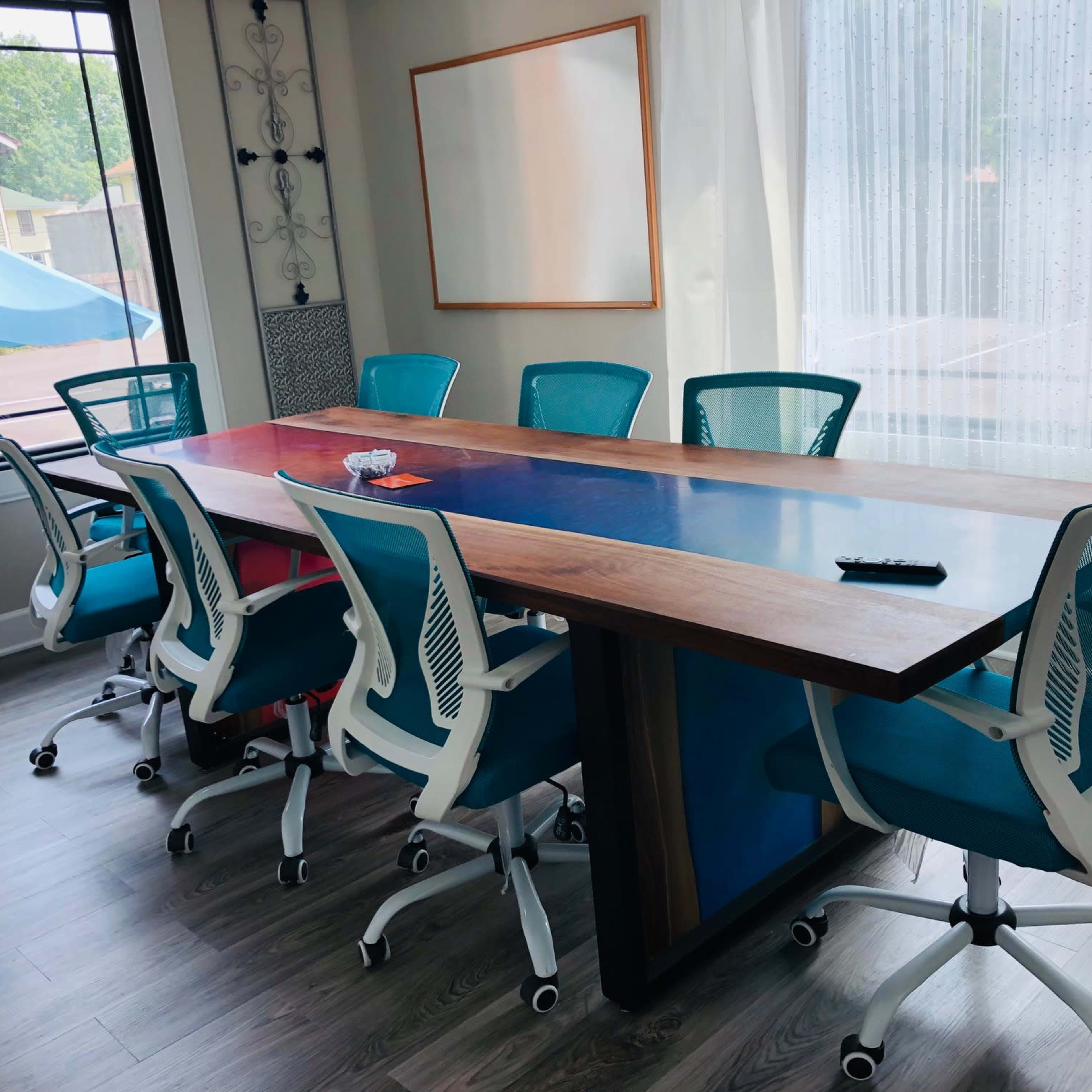 A conference room with a large wooden table and eight blue and white office chairs arranged around it, illuminated by natural light from a window.
