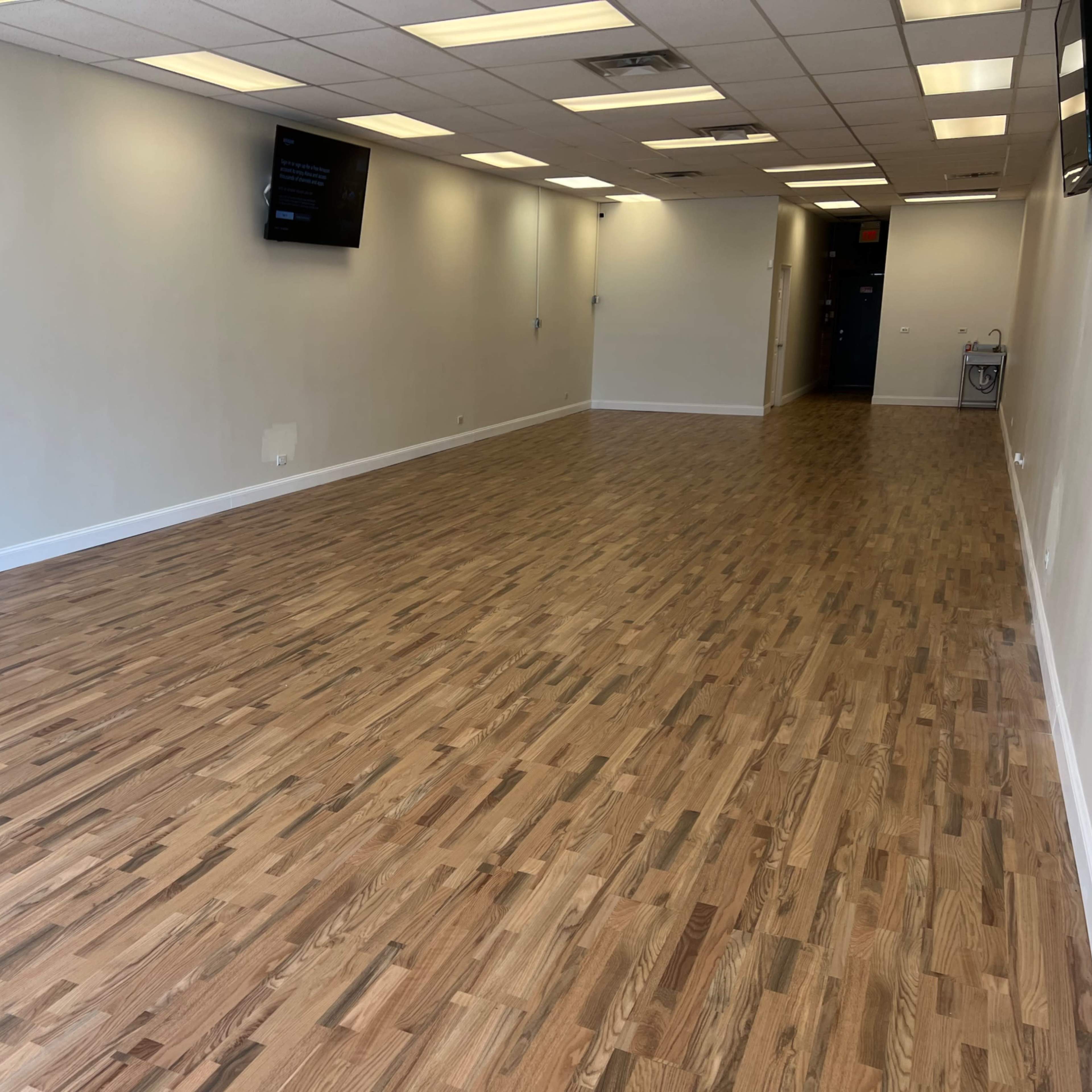 A wide, empty room with light wooden flooring and neutral-colored walls, featuring two wall-mounted screens.
