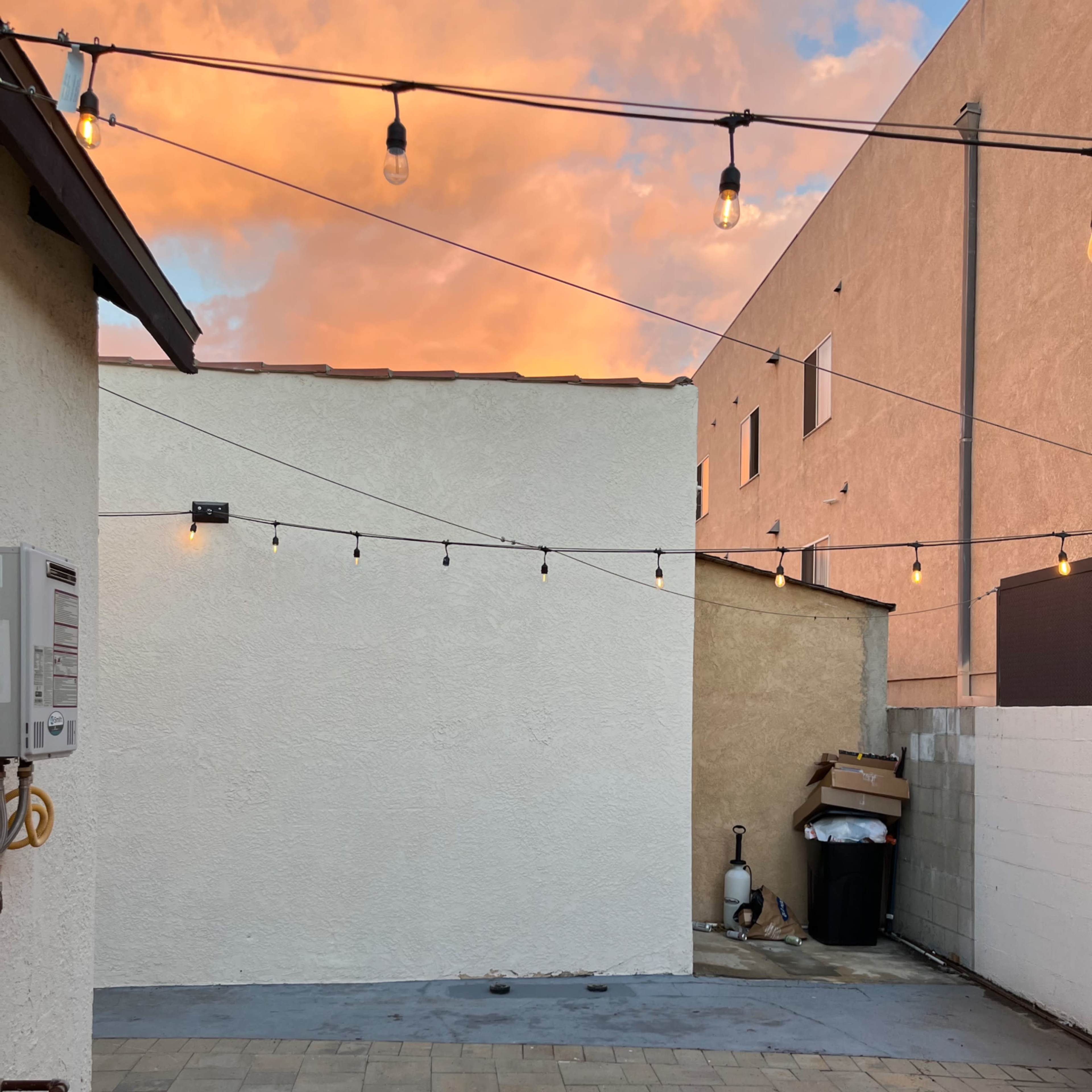 A small outdoor area with paved flooring, a white wall, and string lights overhead, set against a colorful sky at sunset.