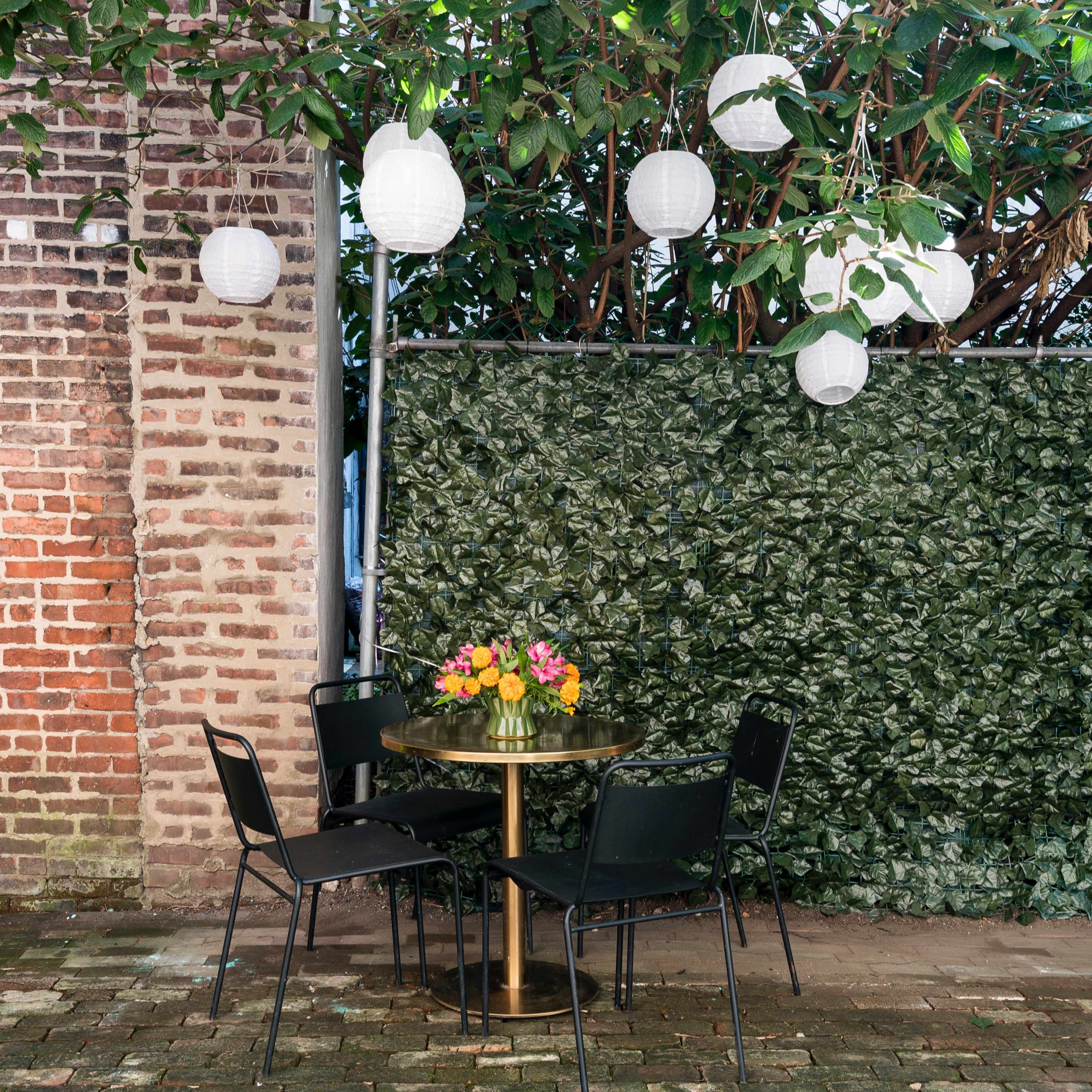 A small round table with a flower arrangement is surrounded by four black chairs beneath hanging lanterns, next to a brick wall and a leafy green backdrop.
