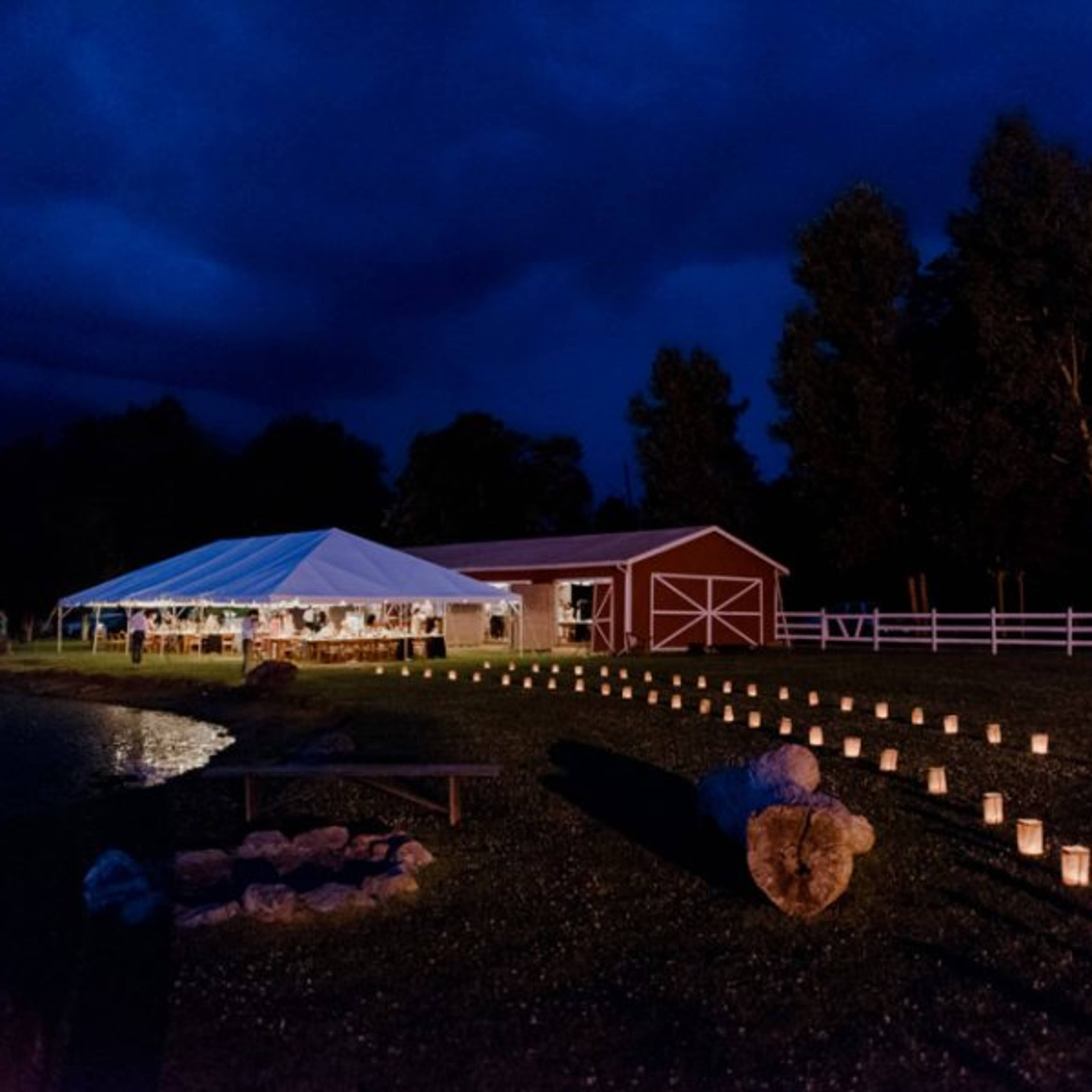 A large tent with lights is set up beside a pond and a red barn, illuminated by lanterns lining a pathway.