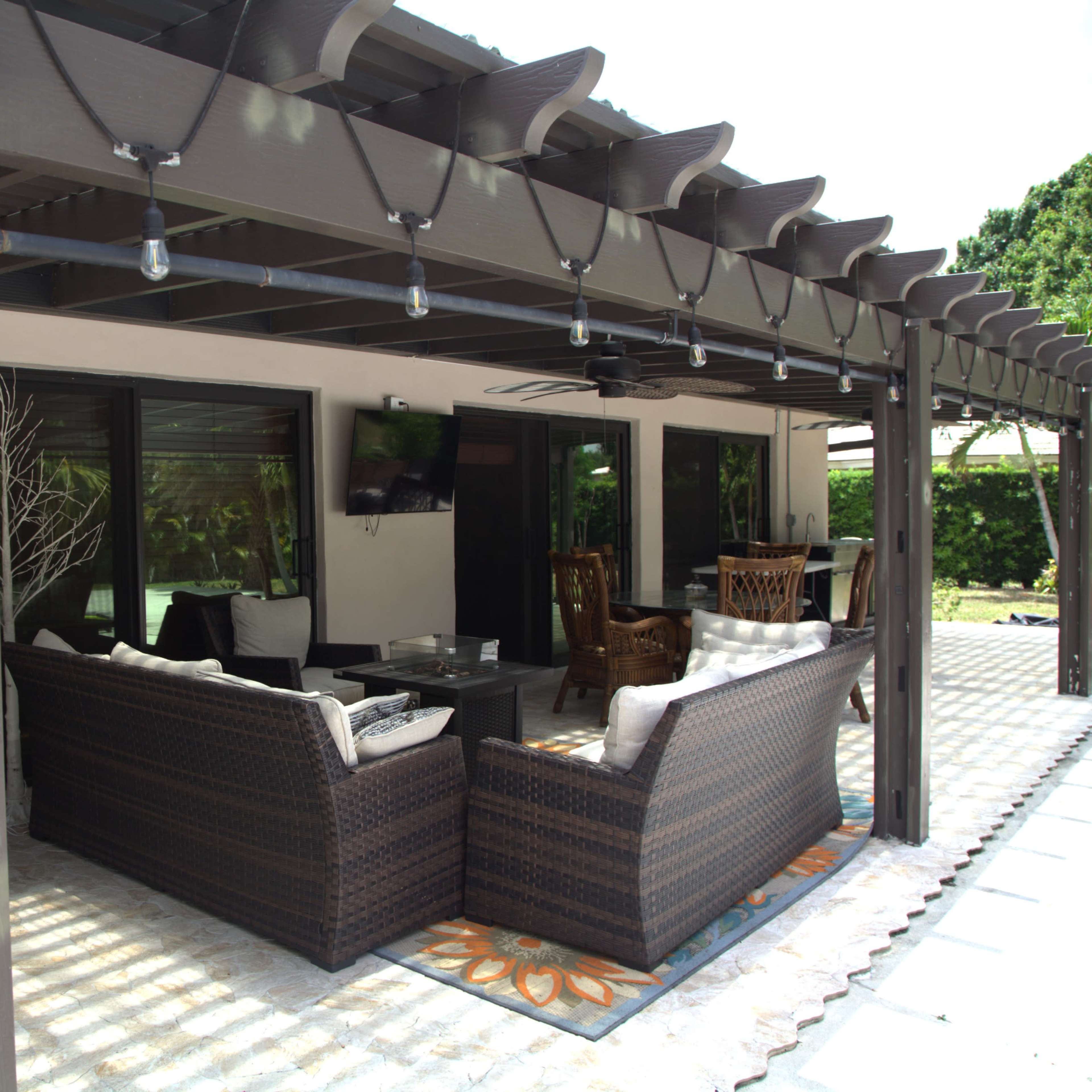 A covered patio with outdoor furniture, including a seating area and dining table, surrounded by greenery.