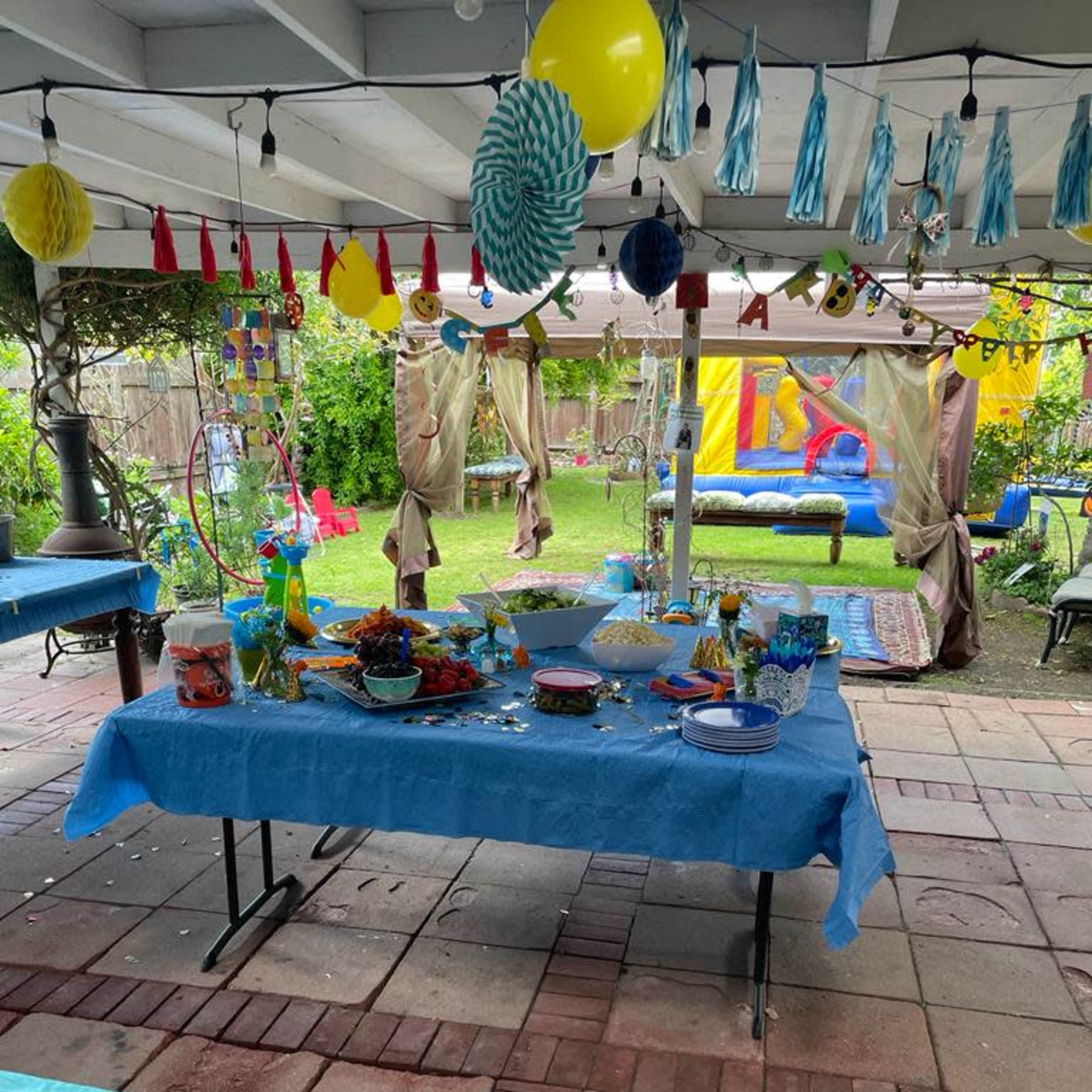 A decorated outdoor party area with tables covered in blue tablecloths, various snacks and decorations, and a colorful bouncy castle in the background.