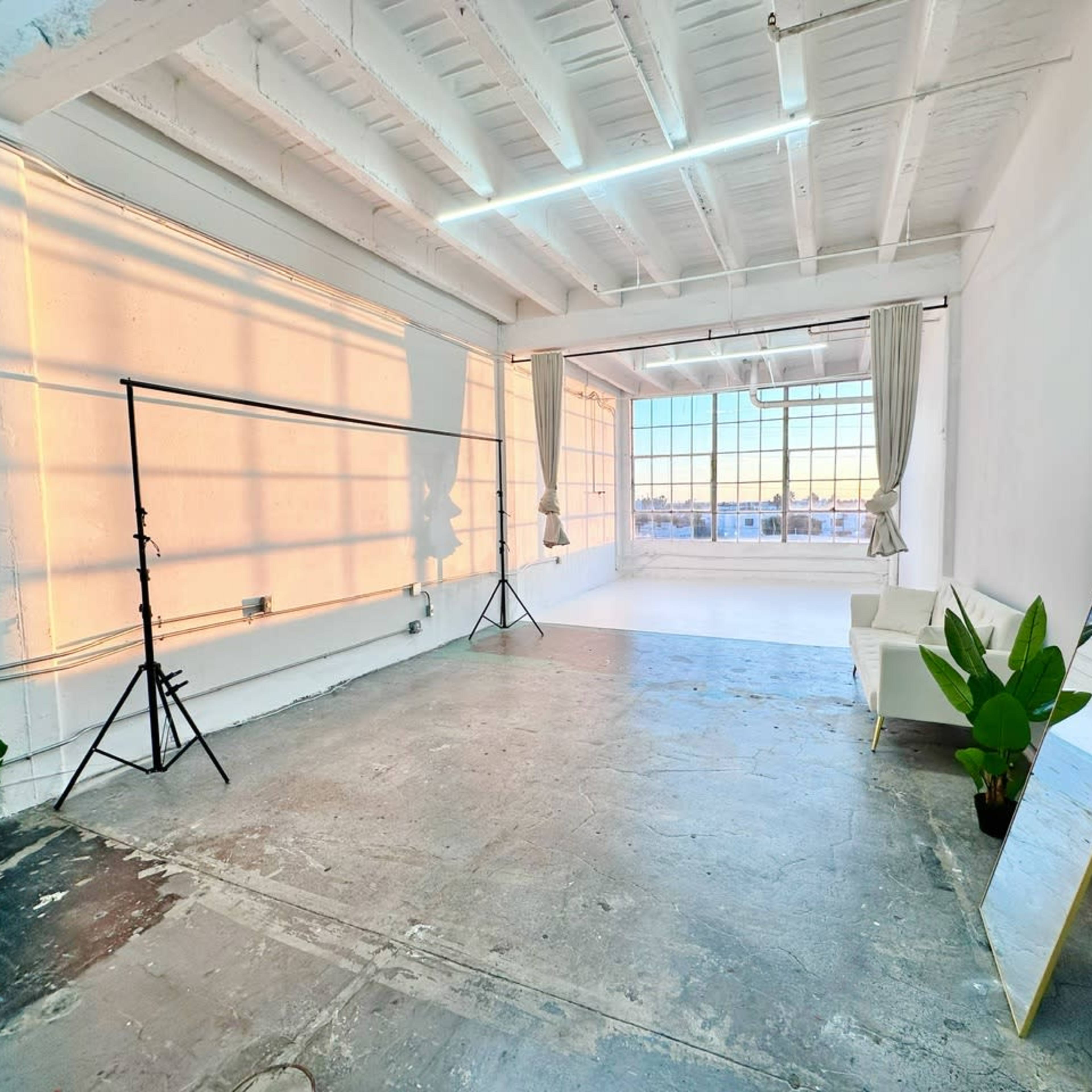 A spacious, brightly lit studio with large windows, a white sofa, a mirror, plants, and lighting equipment set up against a bare wall.