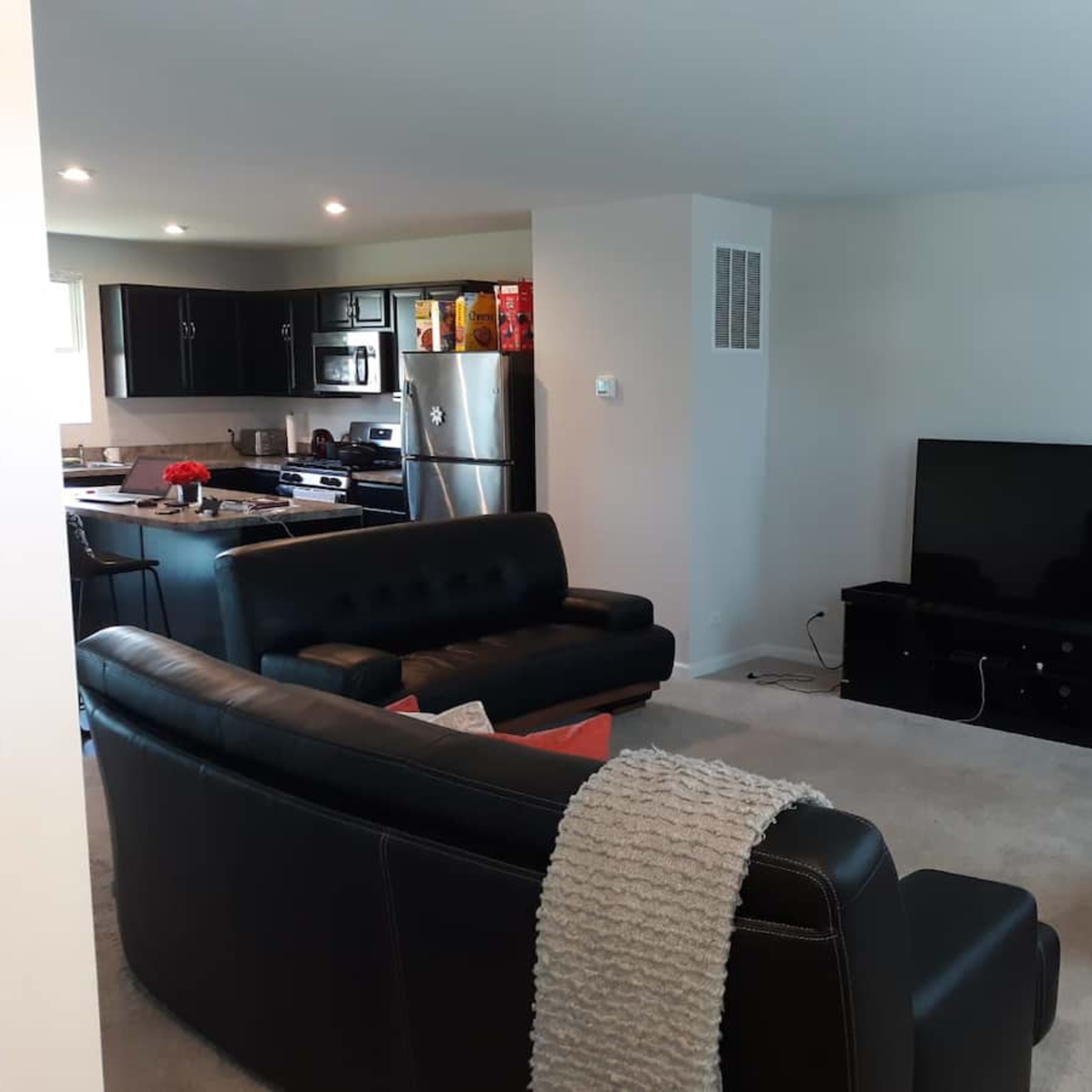 A modern living room with two black sofas, an orange armchair, a television on a stand, and an open kitchen area in the background.