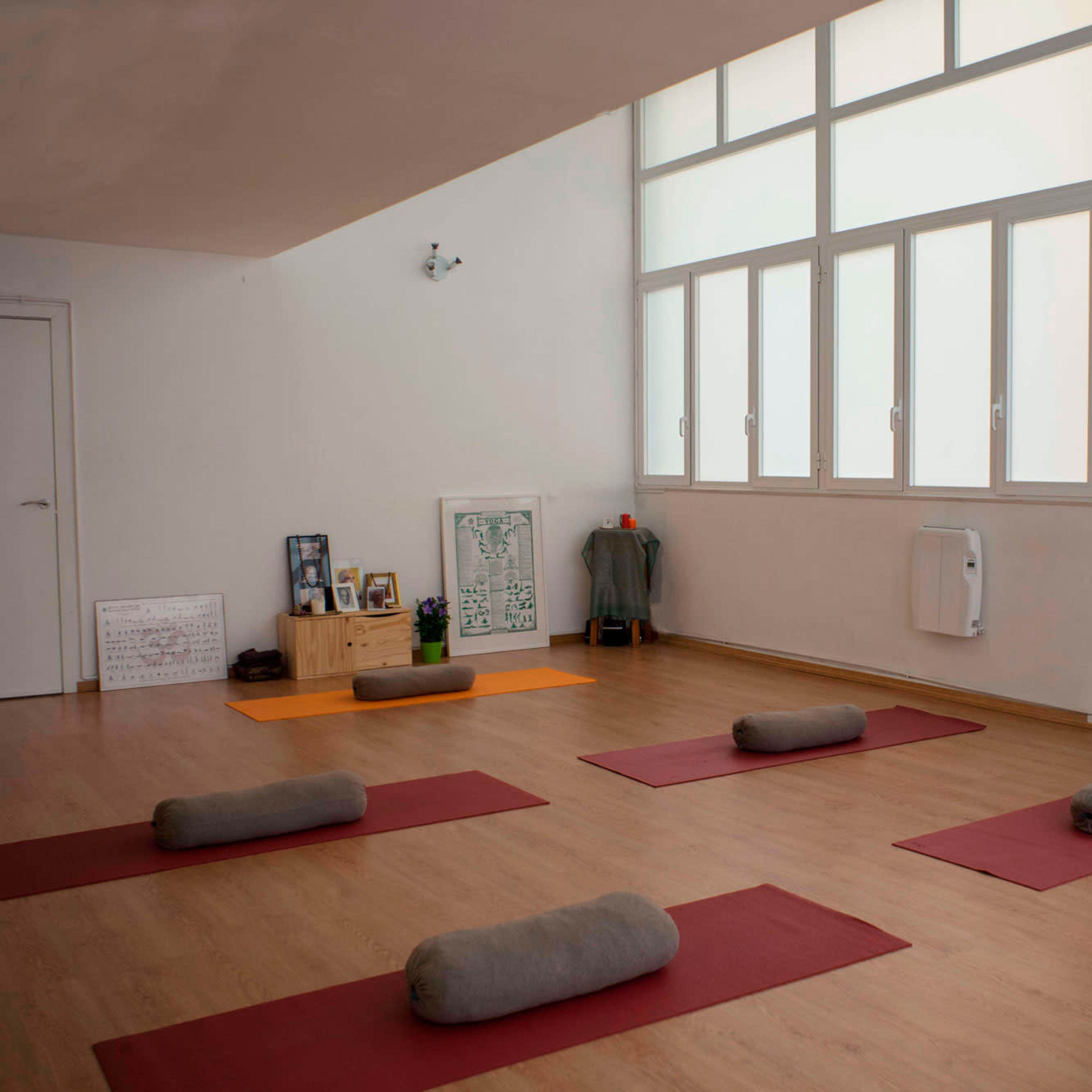 A spacious room with wooden floors, featuring several yoga mats arranged on the floor and large windows allowing natural light to fill the space.