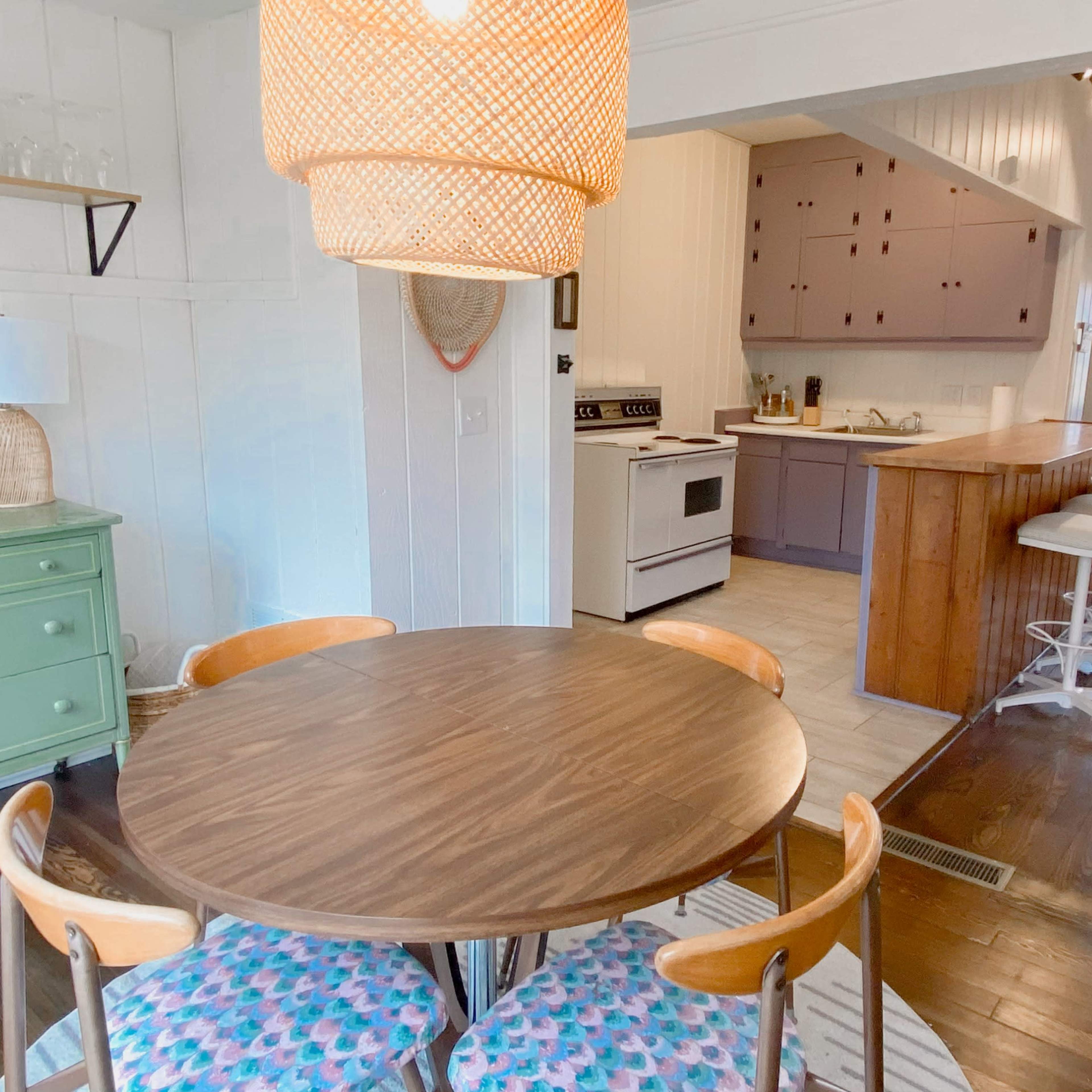 A round wooden dining table with pink and blue patterned chairs is placed in a bright kitchen area featuring a green sideboard and a large pendant light above.