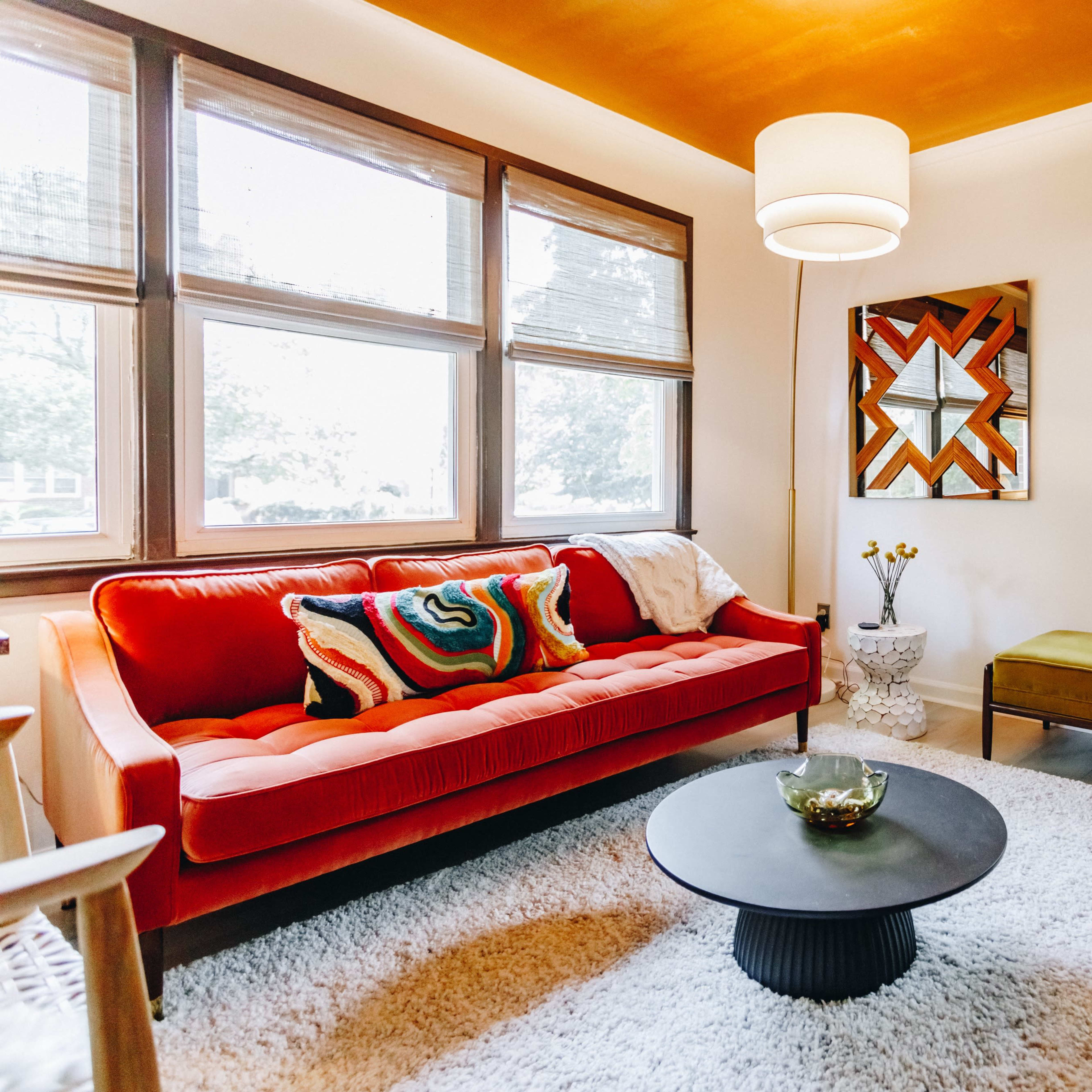 A living room with a red velvet sofa, an olive green bench, a round black coffee table, and large windows allowing natural light to enter.