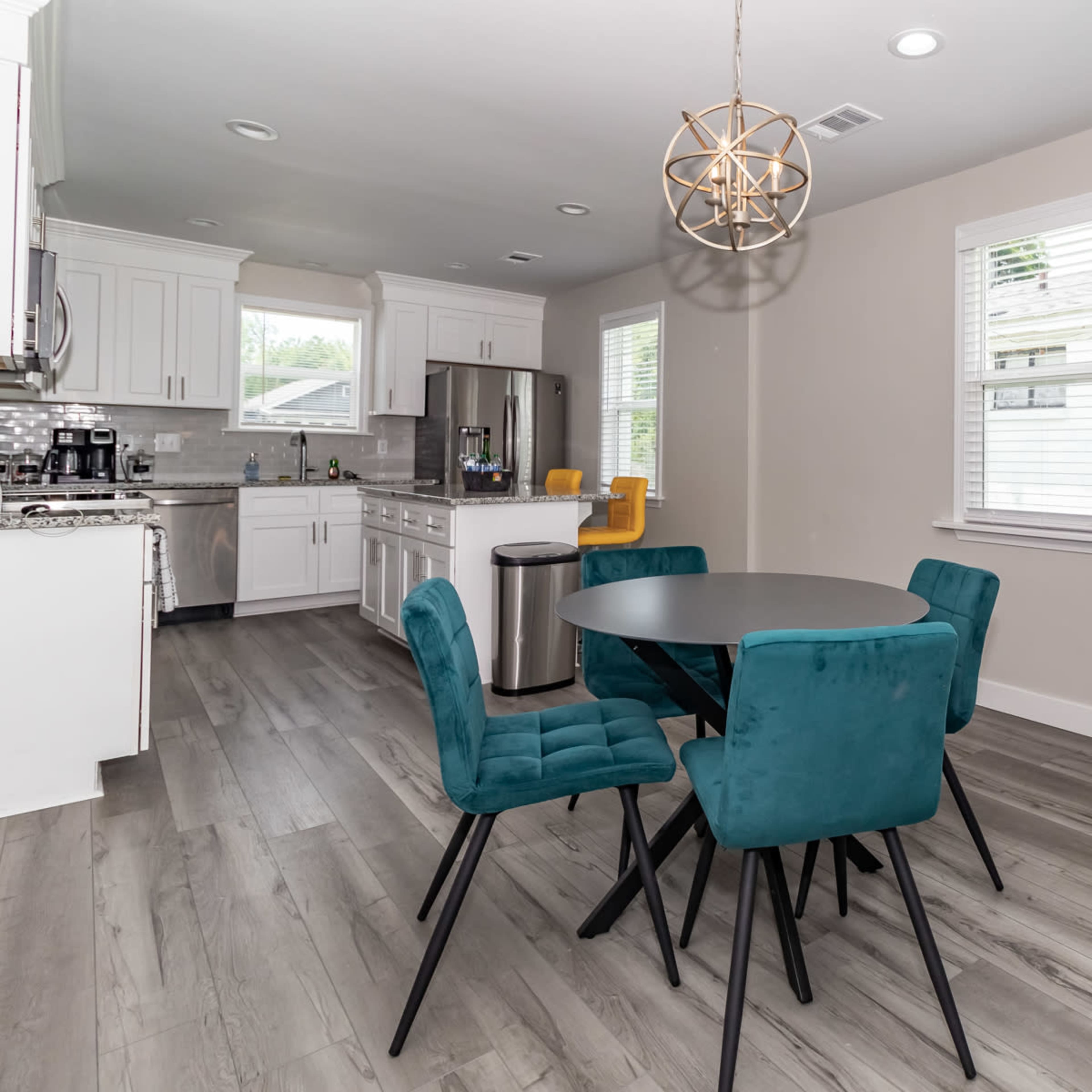 A modern dining area features a round black table surrounded by four blue chairs, with a kitchen visible in the background.