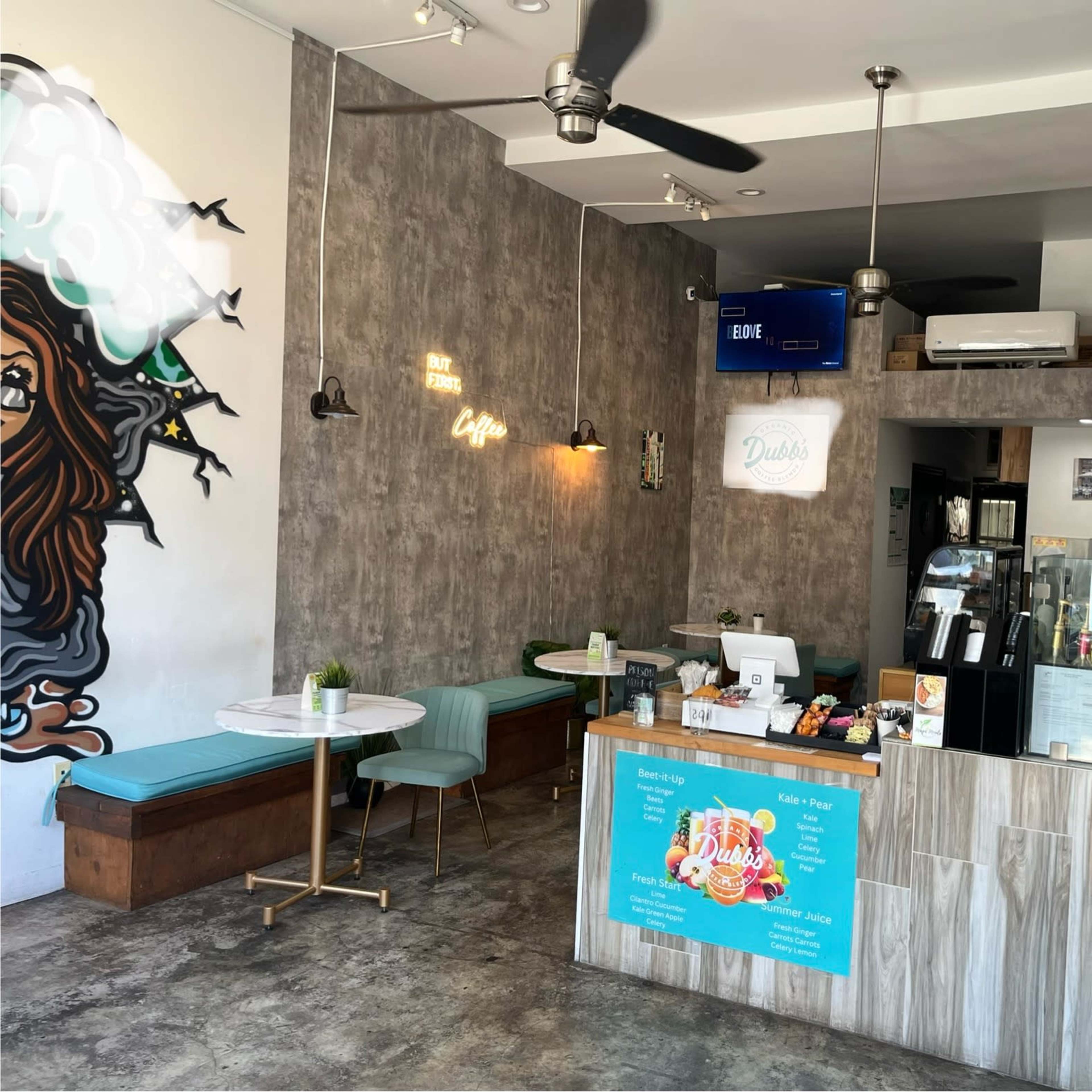 A modern café interior with a serving counter, a wall mural, and seating areas featuring light blue chairs and tables.