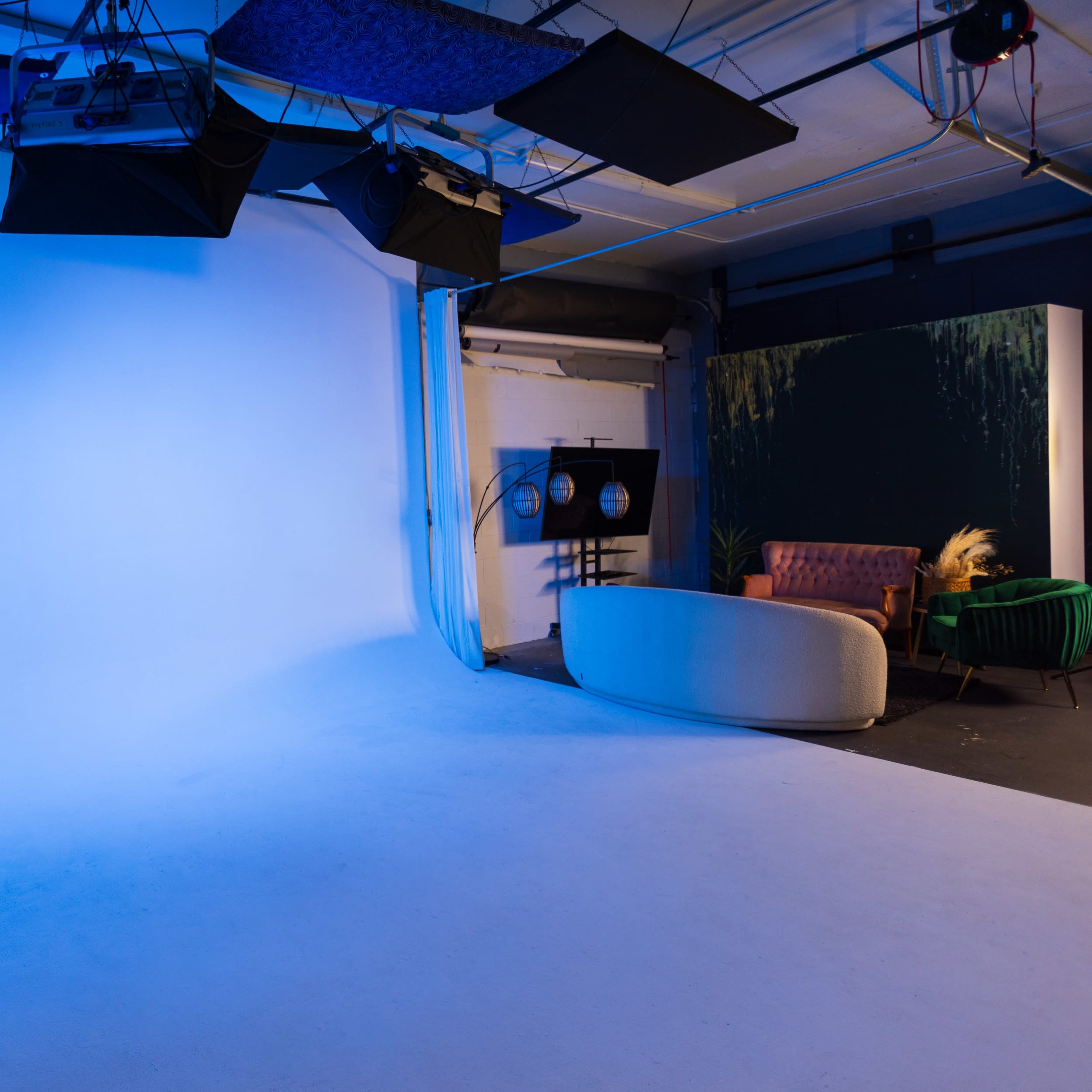 A photo studio with a blue backdrop, lighting equipment, and a minimalistic set featuring a pink sofa and a green armchair.